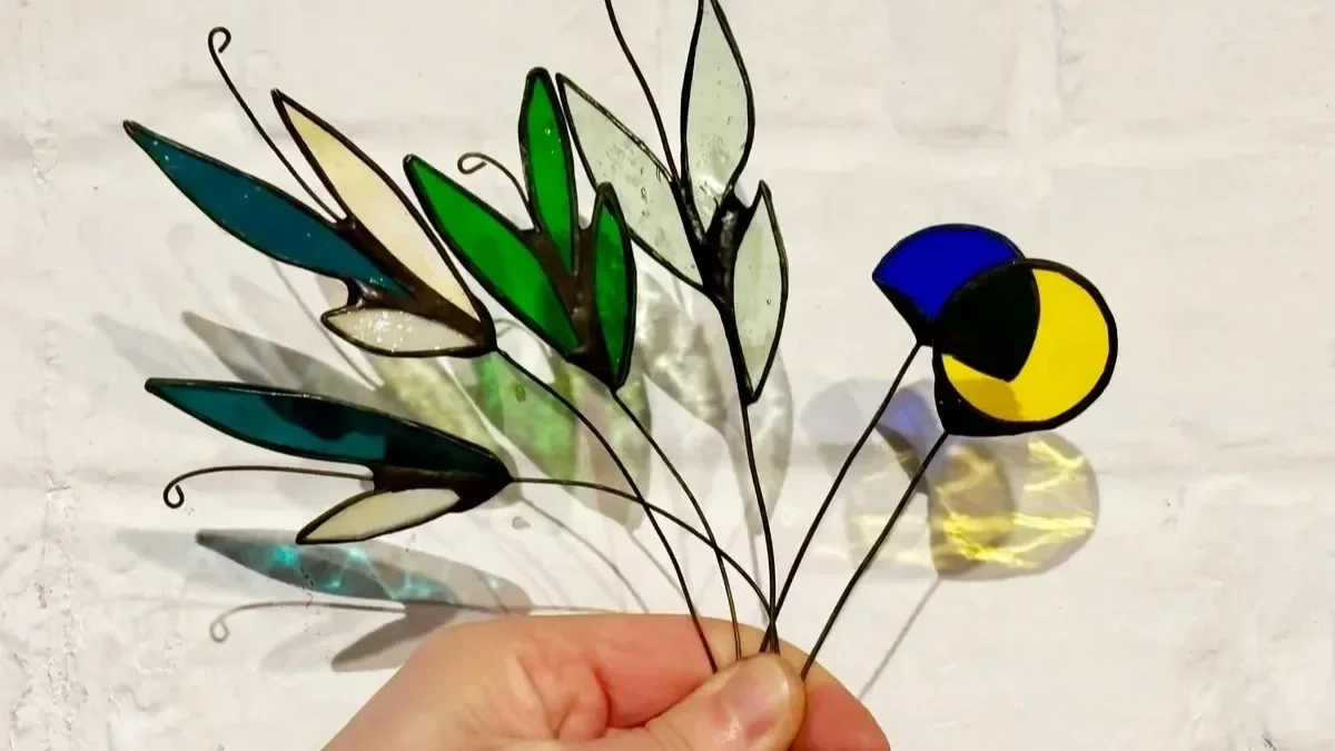 Main tenant un bouquet de fleurs en vitrail coloré créé dans l'atelier de Diane à Lille lors d'un atelier découverte, avec une composition de feuilles vertes et fleurs en verre.