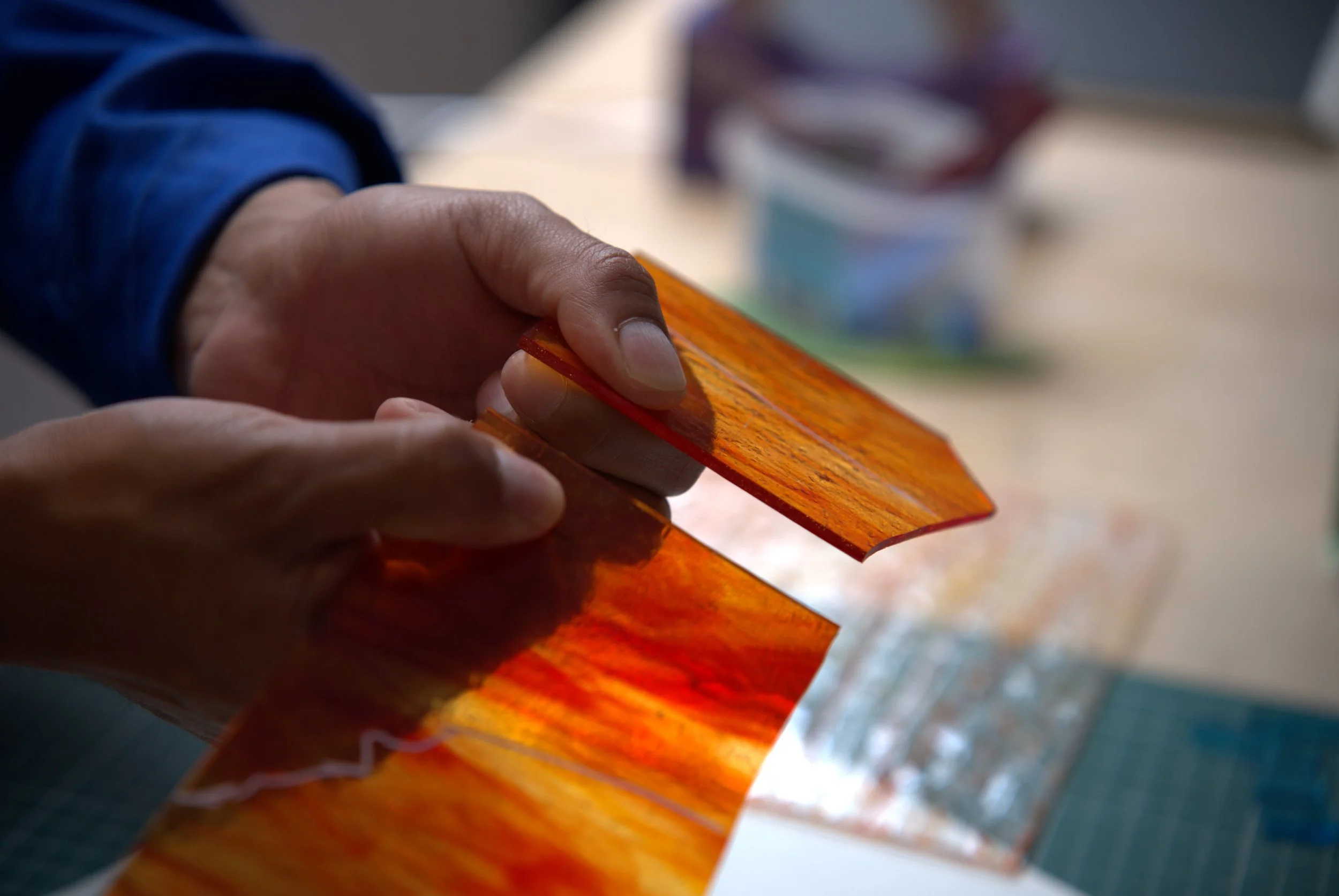 Mains tenant une plaque de verre coloré orange sur un plan de travail pour les vitraux. Une vitrailliste coupe une plaque de verre pour fabriquer un luminaire.