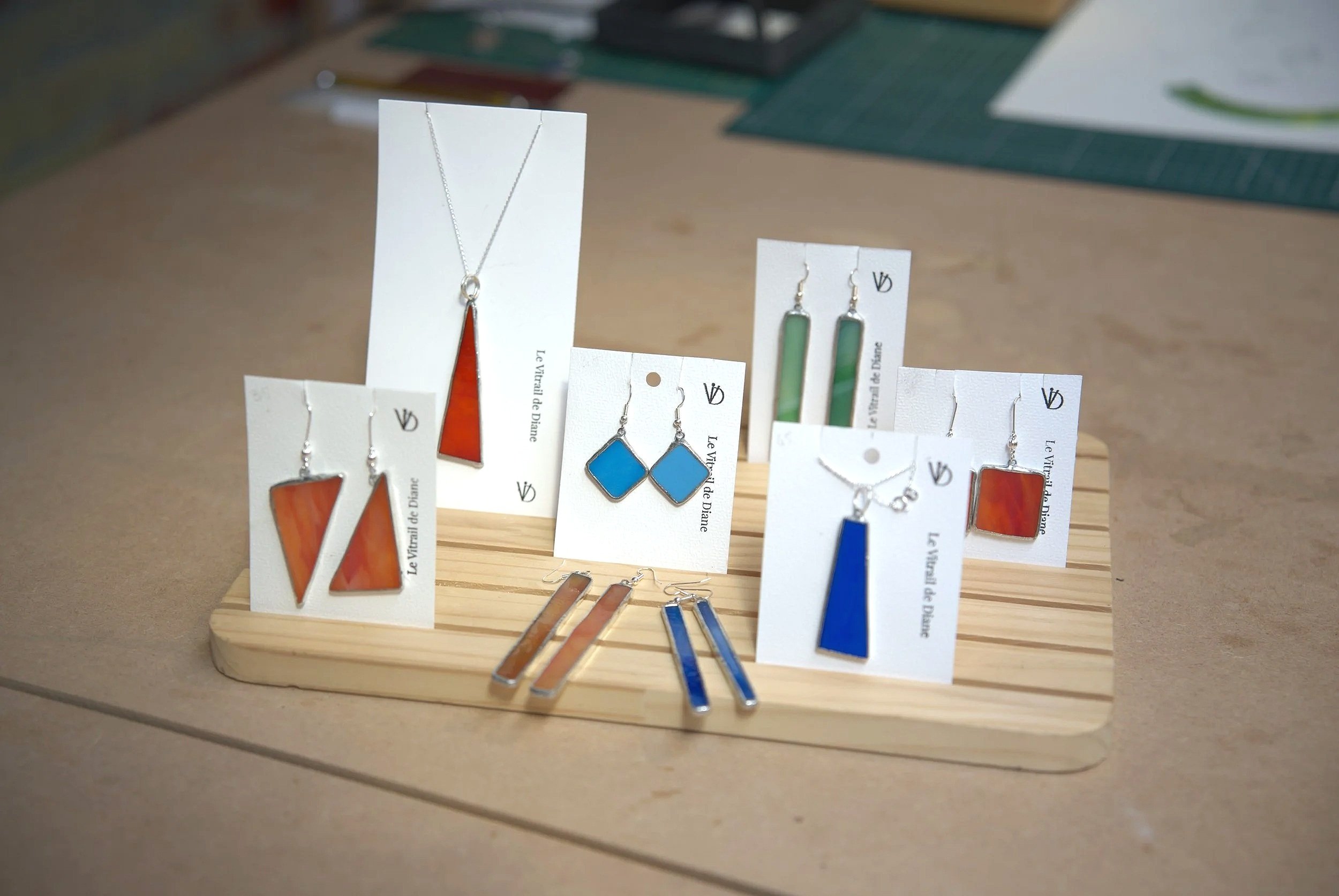 Collection de boucles d'oreilles et pendentifs colorés en vente, présentée sur une plateforme en bois, avec des étiquettes portant le texte "Le Vitrail de Diane".