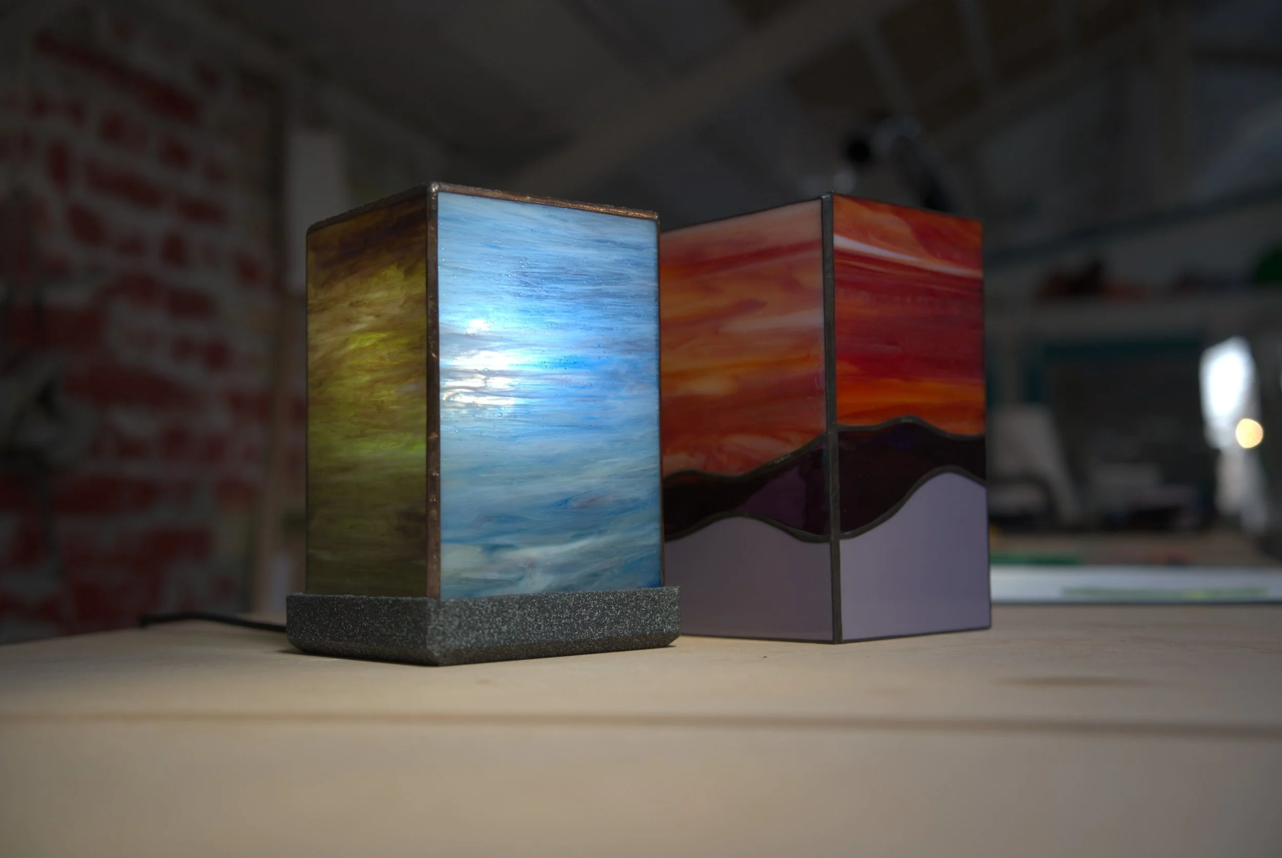 Deux lampes en verre peint, une avec un motif de ciel bleu et l'autre avec un motif de coucher de soleil, posées sur une table en bois.