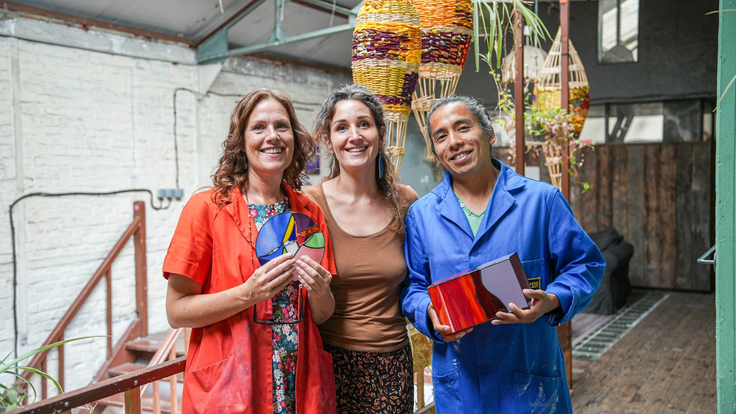 Trois personnes dans un atelier, tenant des objets en verre coloré, souriant à la caméra, décorations en osier suspendues en arrière-plan. Les participants de l'ateliers vitrail sont ravis de leurs créations en vitrail.