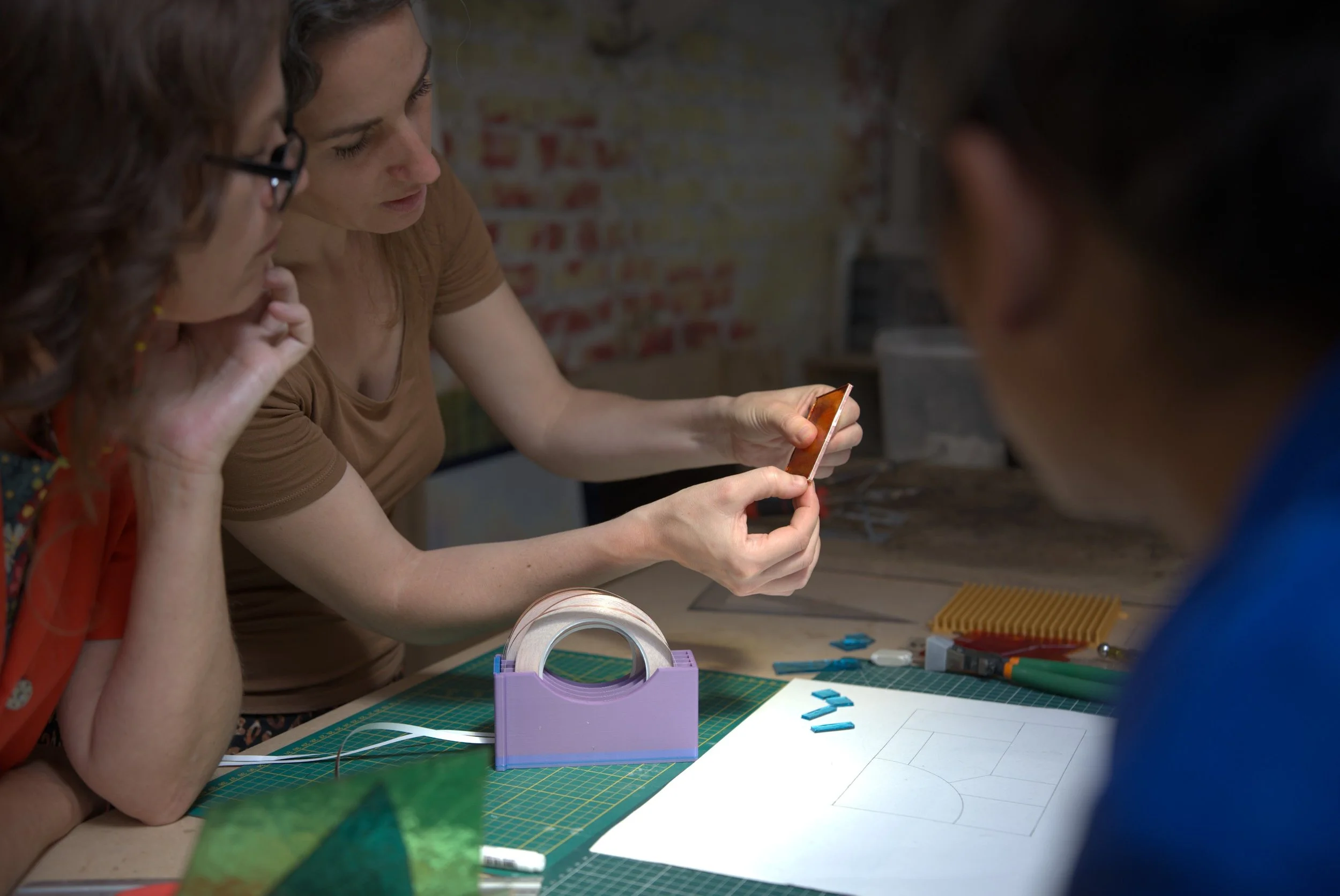 Trois femmes discutent autour d'une table de travail avec divers outils, une d'elles montre un téléphone portable, un plan de terrain dessiné est posé sur la table.