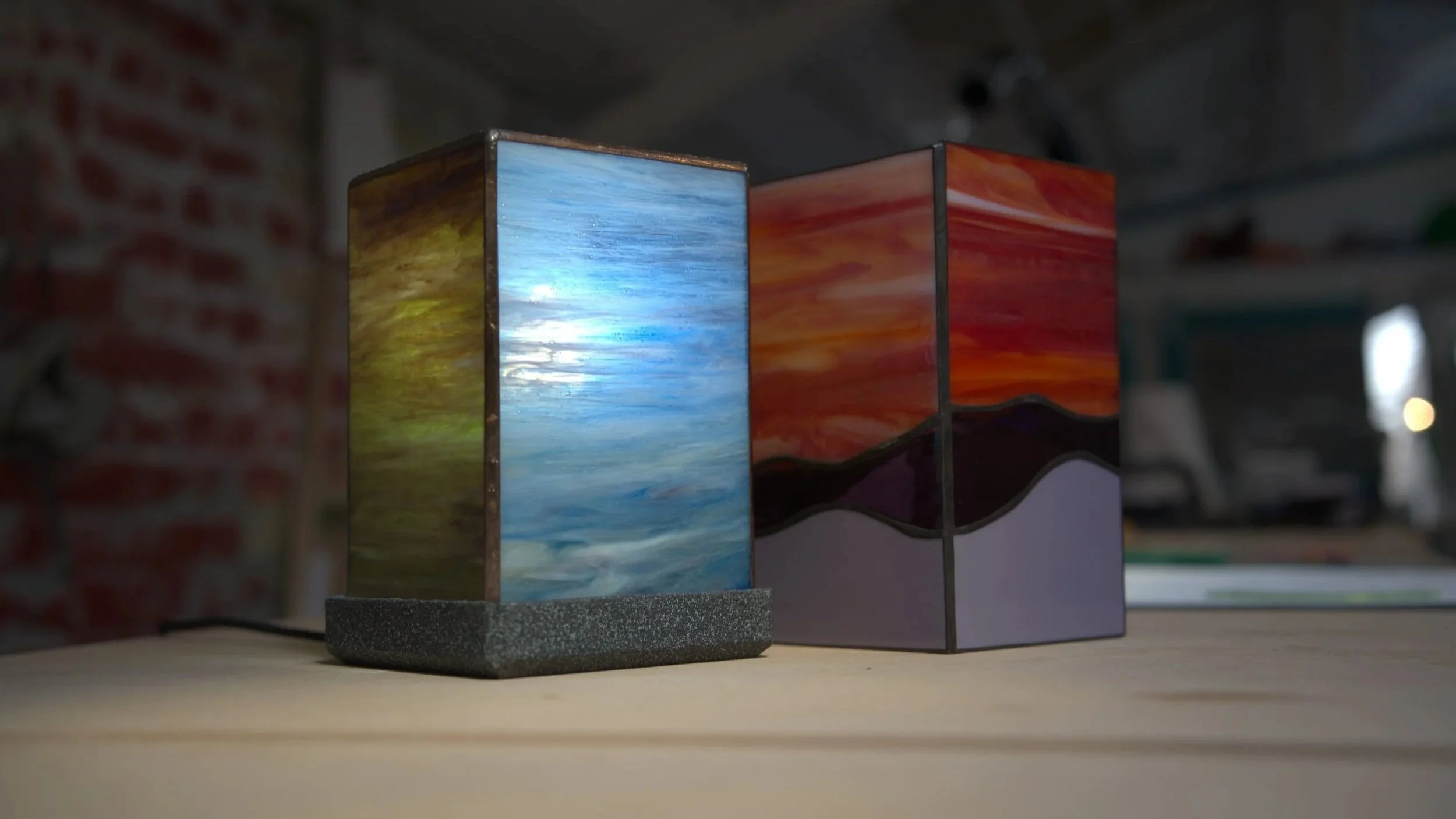 Deux lampes en bois peint avec des paysages impressionnistes, une nature et un coucher de soleil, placées sur une surface en bois. lors d'un atelier création en vitrail à Lille dans l'atelier de Diane à Lille.