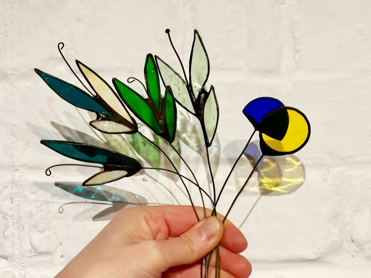 Main qui tient un ensemble d'abats en verre coloré en vitrail fabriqué lors d'un atelier créatif dans l'atelier de Diane à Lille,  représentant des feuilles et de s fleurs , avec des ombres projetées sur un mur blanc.