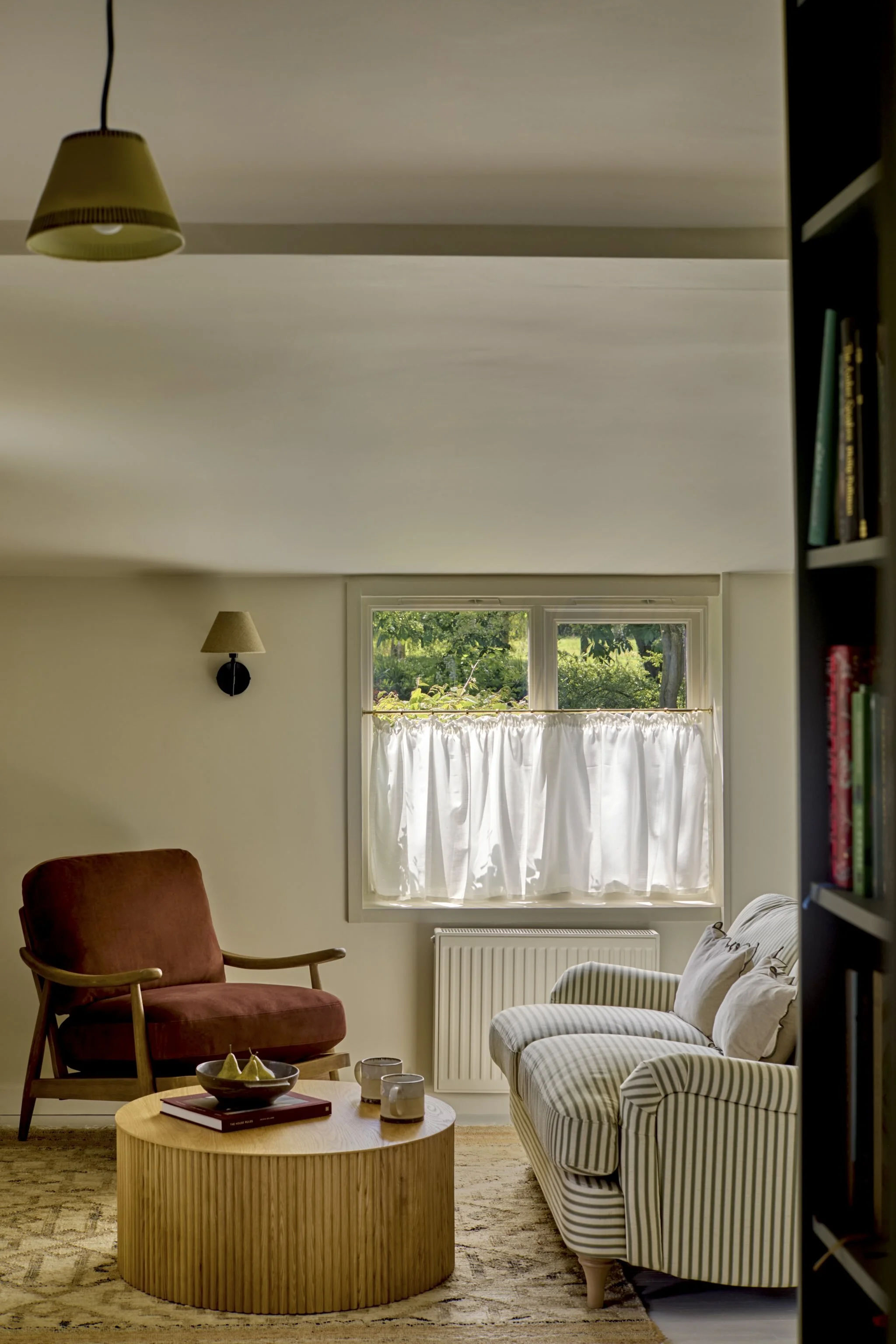 Living room with striped armchair, orange armchair, window with white curtains, bookshelf, and wall lamps.