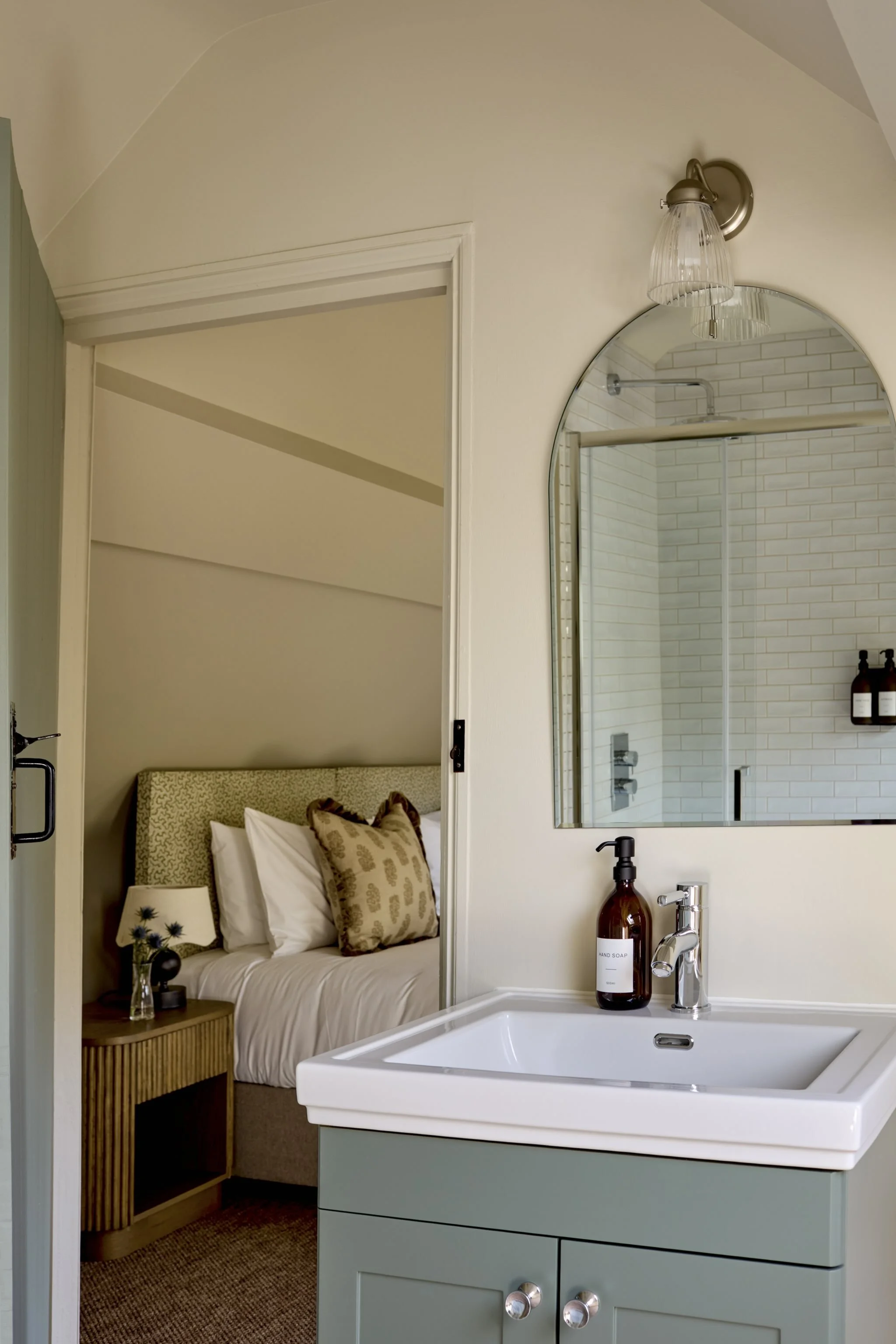 A cozy bedroom with a bed, pillows, a bedside table with a lamp, a bathroom mirror, a shower with white subway tiles, and a sink with a soap dispenser.