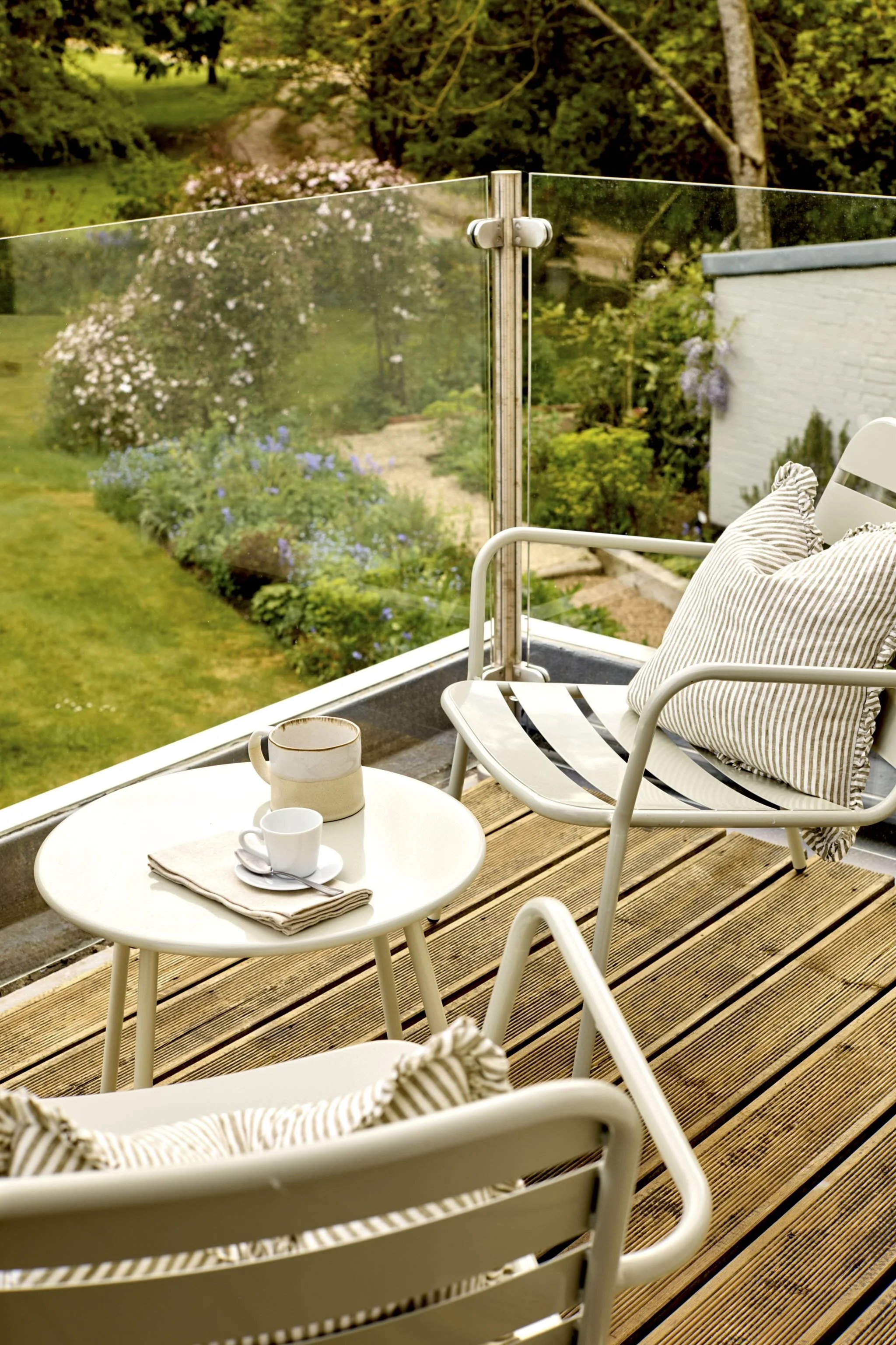 A patio with a white table and chairs, with coffee cups and a napkin, overlooking a garden with trees and colorful flowers, enclosed by a glass railing.