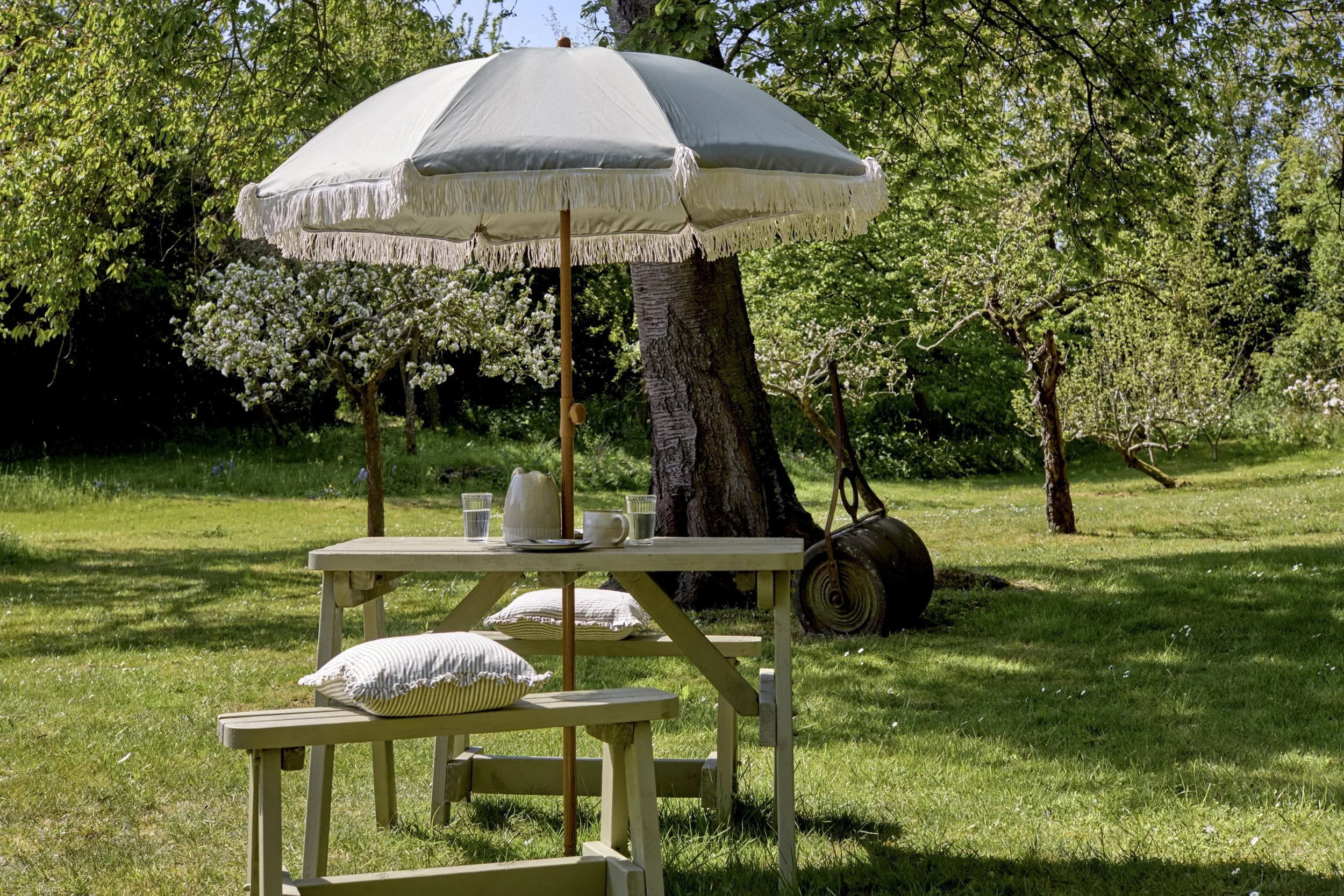 Outdoor patio scene with a white wooden table and cushions, a large white parasol with fringe, surrounded by green trees and grass.