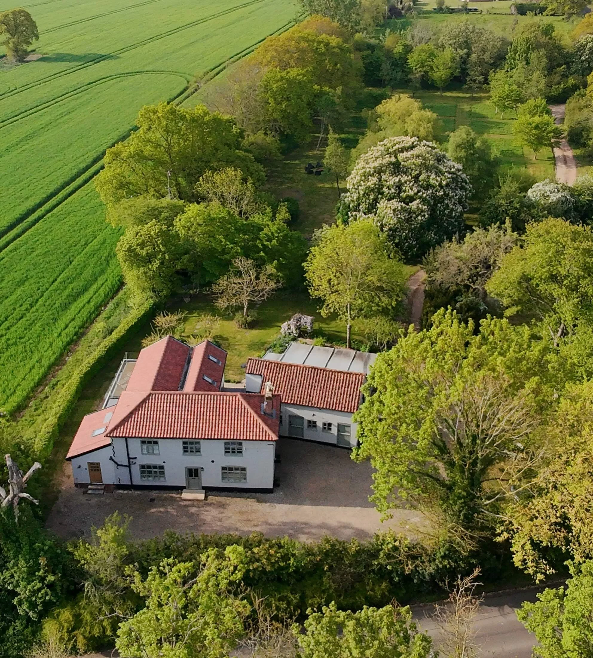 Aerial view of a large white house with a red tile roof surrounded by trees, greenery, and a gravel driveway in a rural area.