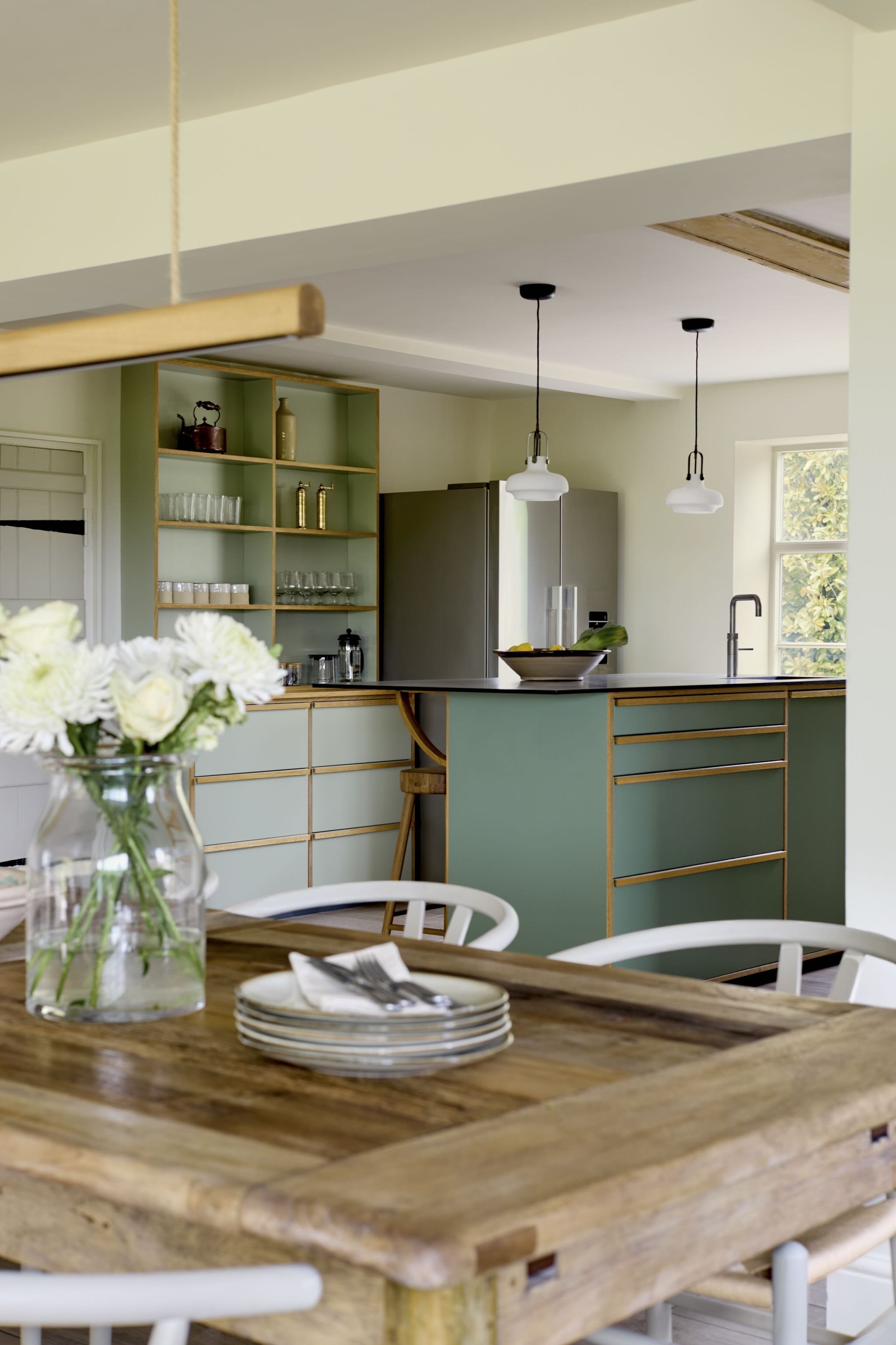 Open kitchen and dining area with a wooden table, white chairs, a green and wood kitchen island, an open shelving unit with glassware and decorative items, pendant lights, and a window with plants outside.