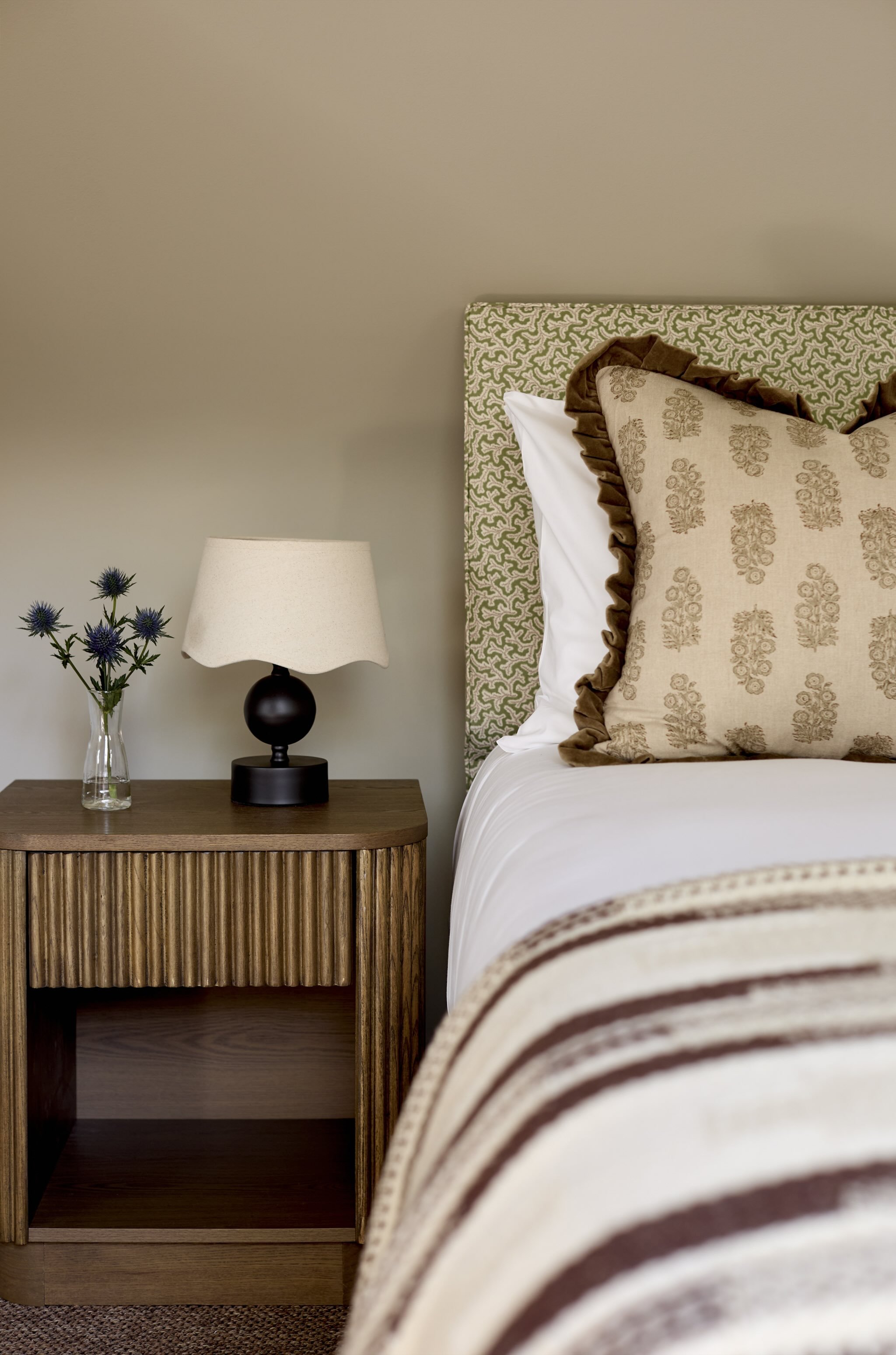 A bedroom with a beige wall, a wooden nightstand with a black lamp and a glass vase with purple flowers, a bed with a green and white patterned headboard, a white pillow, and a decorative pillow with a floral pattern and ruffled brown border.