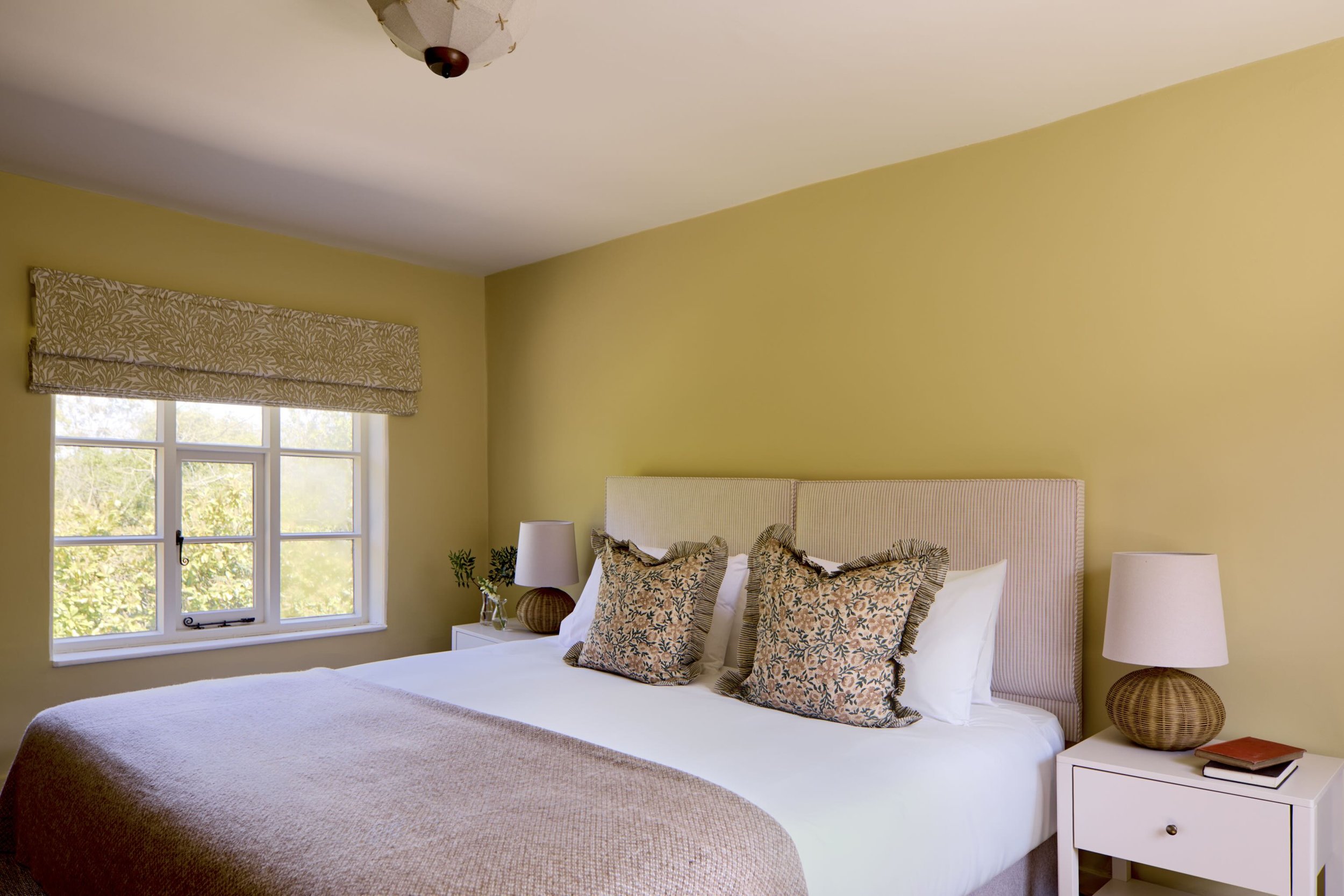 A bedroom with a bed, floral-patterned pillows, white nightstands, a lamp, a window with a patterned valance, and a yellow wall.