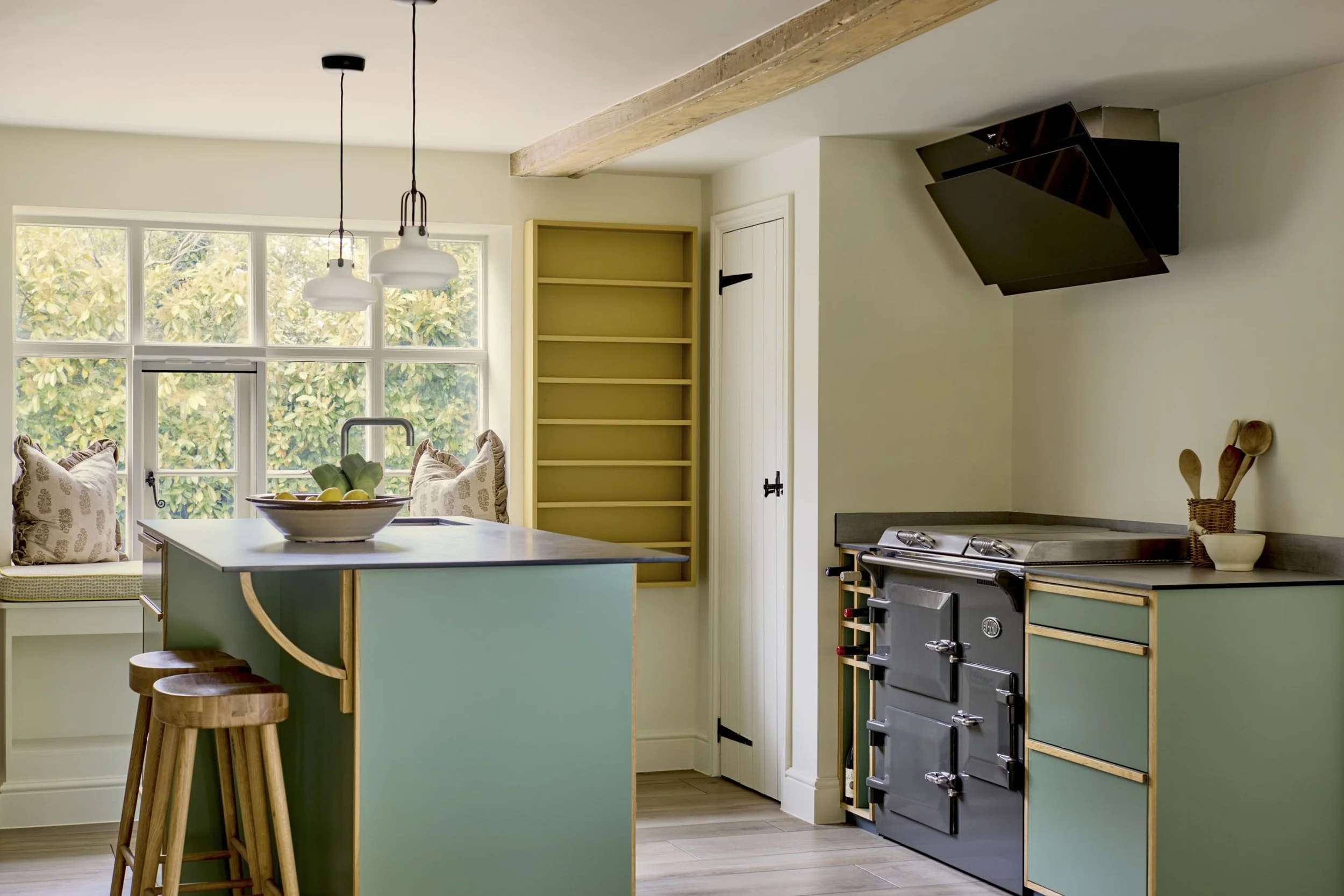 A cozy kitchen with a view of a sitting nook by a large window, featuring a green island with two wooden stools, yellow open shelves, and a black stove with a matching black range hood. Pendant lights hang over the island, and there is a bowl of lemo