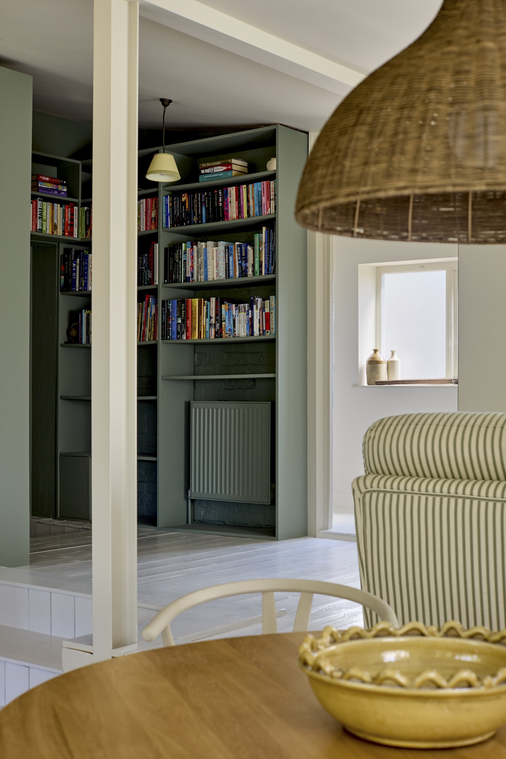 A wooden dining table with a yellow bowl with a decorative edge, in front of a striped armchair. In the background, part of a green bookshelf with books and a window with decorative pottery.