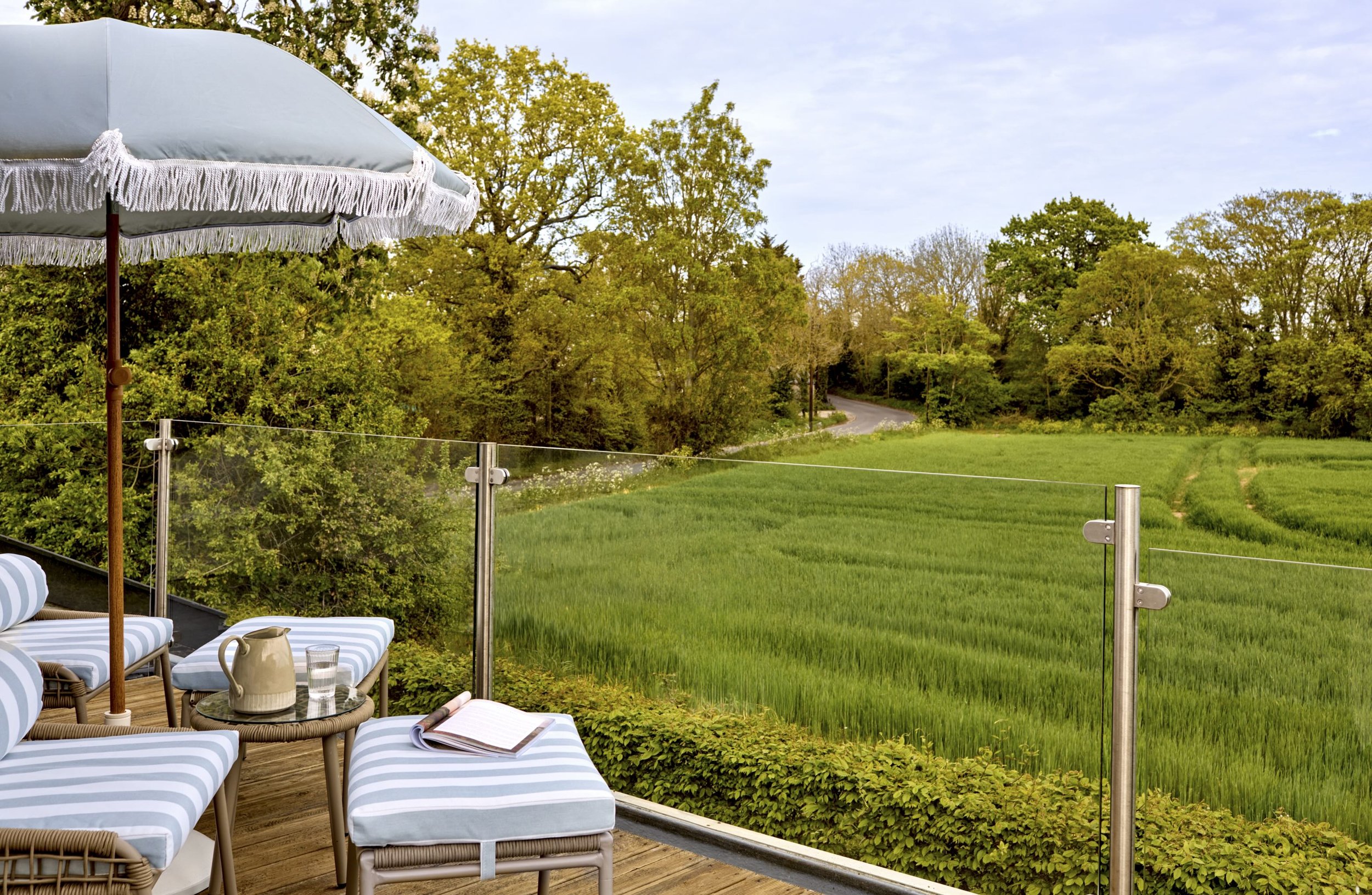 A patio with lounge chairs, a small round table with a pitcher and a glass, a large umbrella, and a view of lush green trees and grass beyond a glass railing.