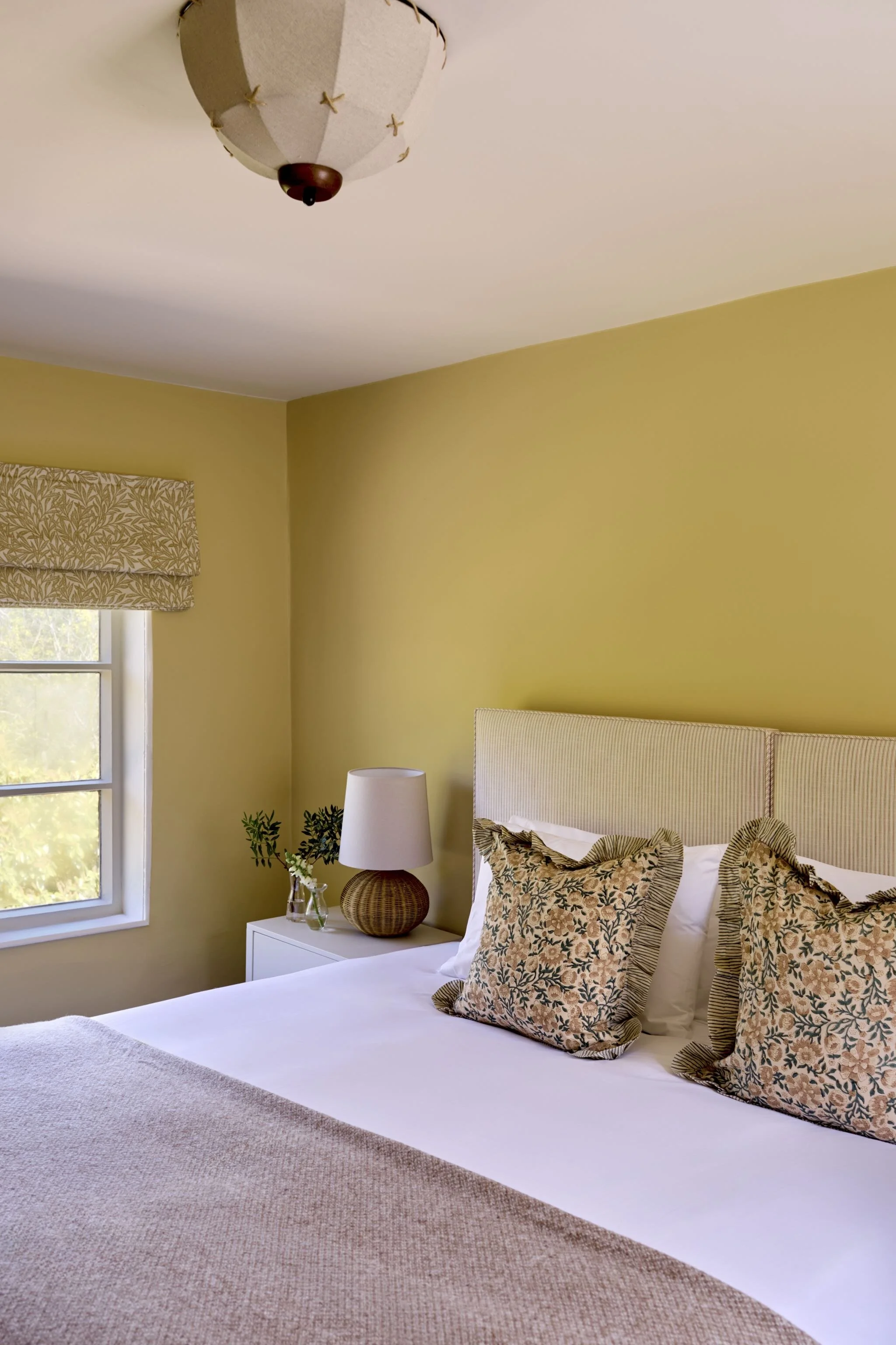 A bedroom with a yellow wall, a window with a patterned valance, a white bedside table with a glass vase and a wicker lamp, a bed with floral pillows, and a beige upholstered headboard.