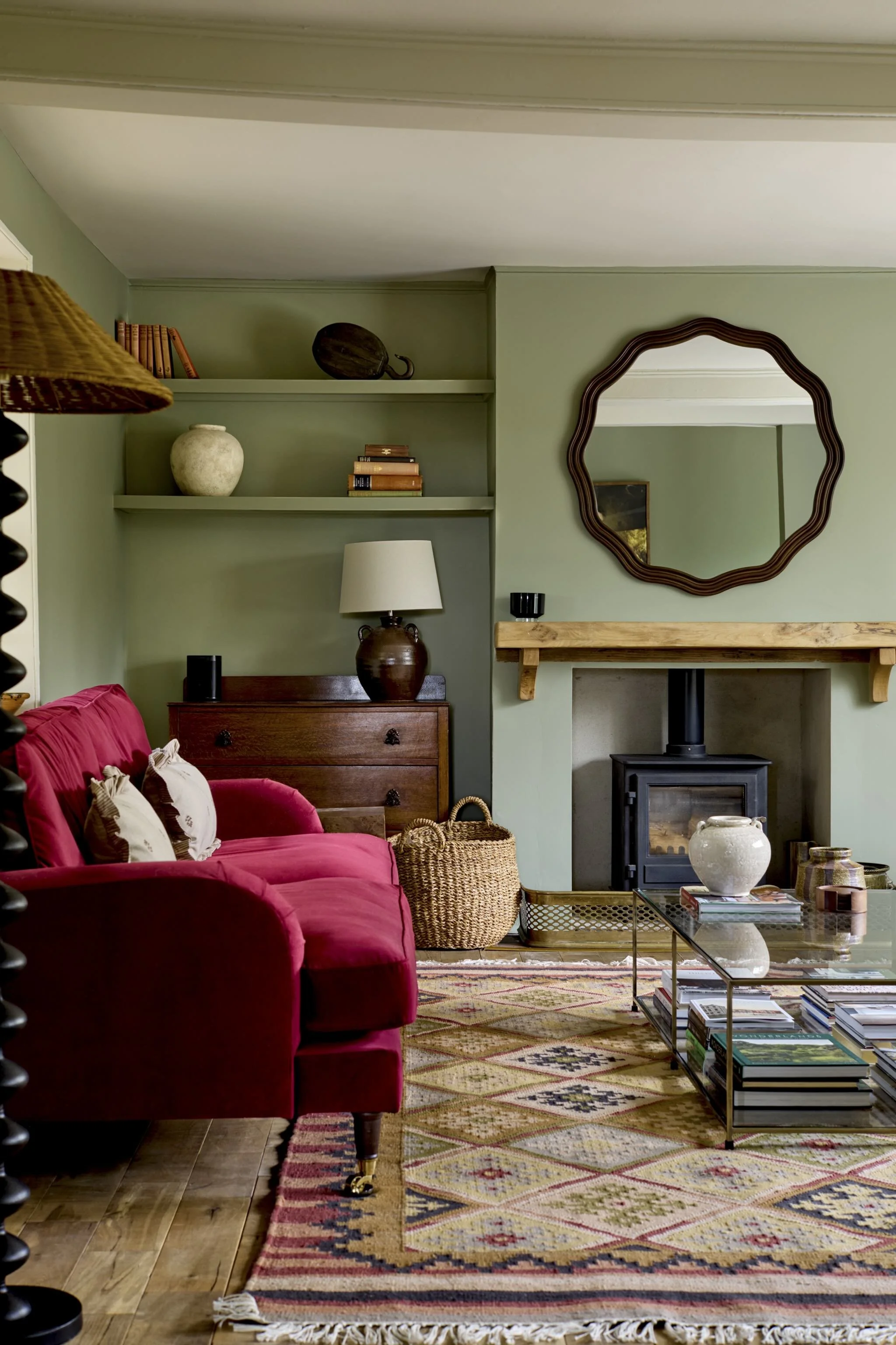 Living room area with a red couch, a glass coffee table with magazines and decorative items, a wood stove, a fireplace with a wooden mantel, a mirror on the wall, a table lamp on a wooden dresser, woven baskets, and a patterned area rug.