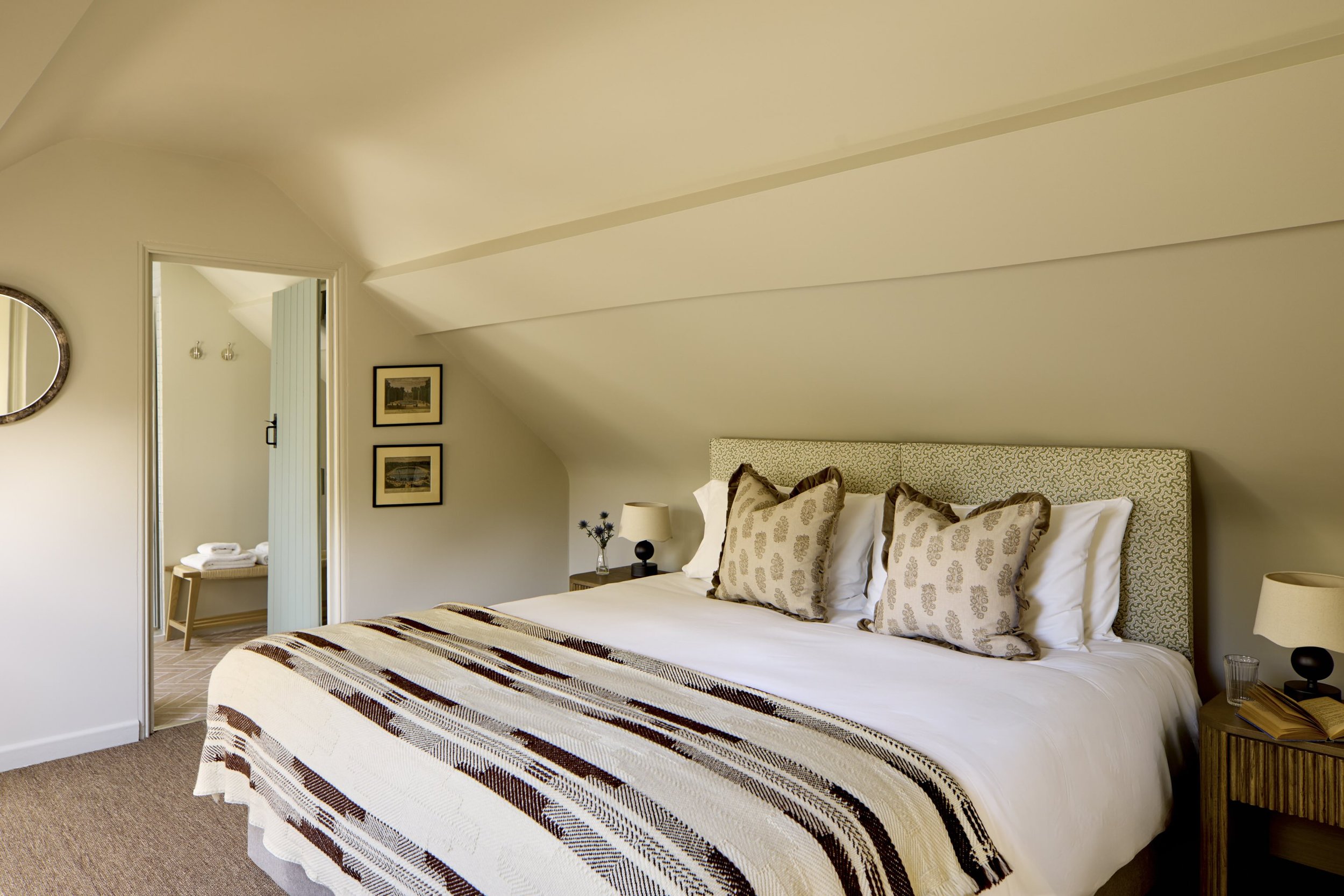 Cozy bedroom with a bed, multiple pillows, nightstand, lamp, and framed pictures on the wall. An open door reveals a small bathroom with towels and hooks.