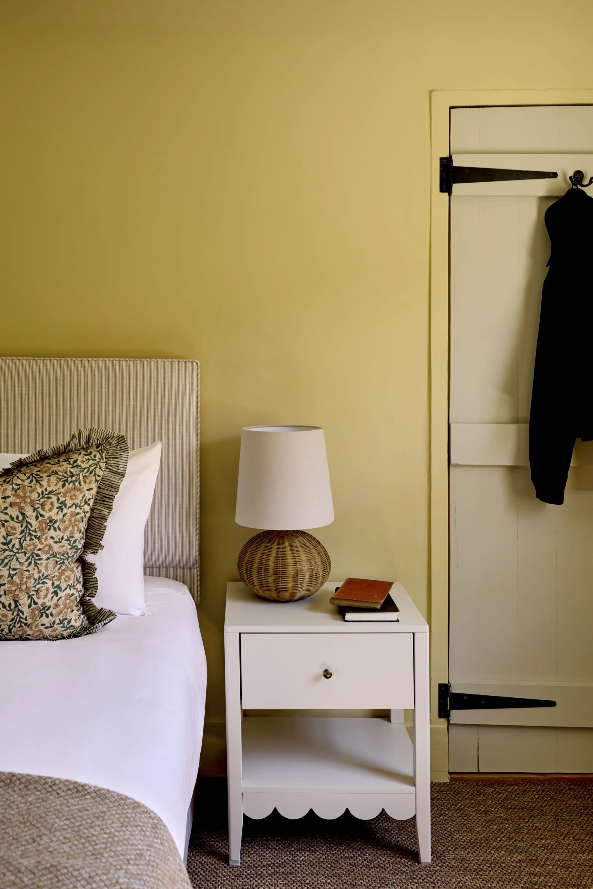 A bedroom with a white bed, a striped beige armchair, a white nightstand with a wicker table lamp and books, a pale yellow wall, and a white closet door with black hardware.