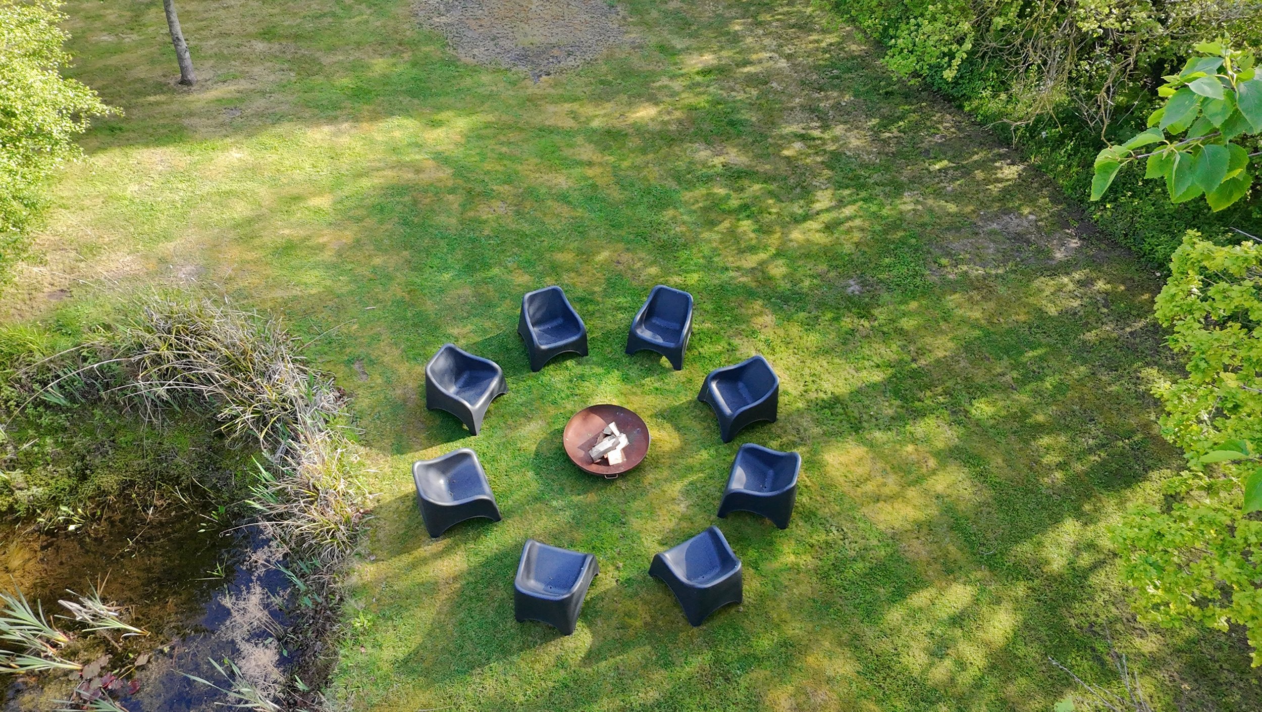 An aerial view of a backyard with green grass, trees, and shrubbery. In the center, there are several black outdoor chairs arranged in a circle around a fire pit.