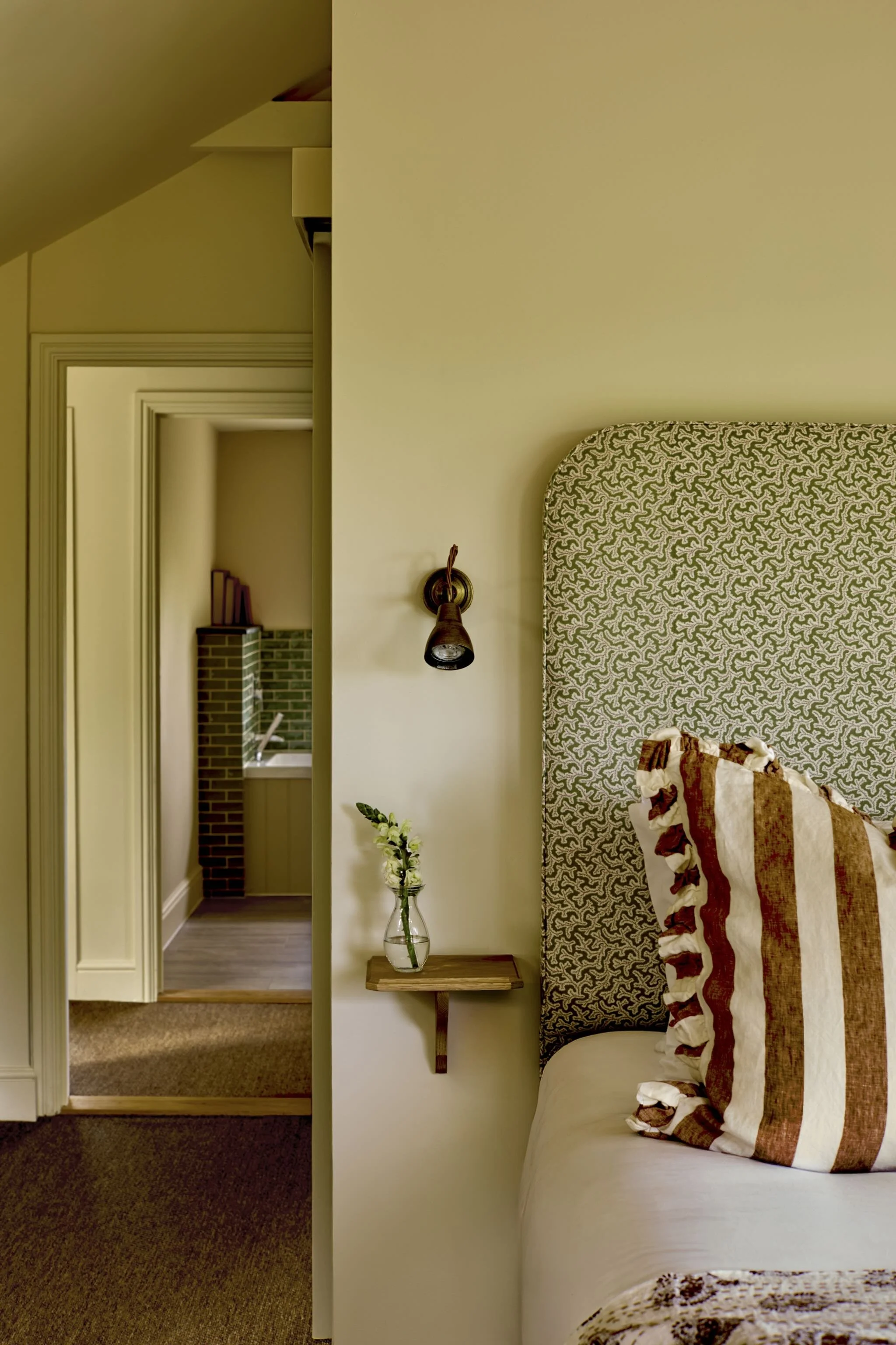 A view from a bedroom showing a part of the bed with a striped blanket, a small wooden side table with a glass vase of flowers, a wall-mounted lamp, and a door leading to a hallway with a fireplace and books in the background.