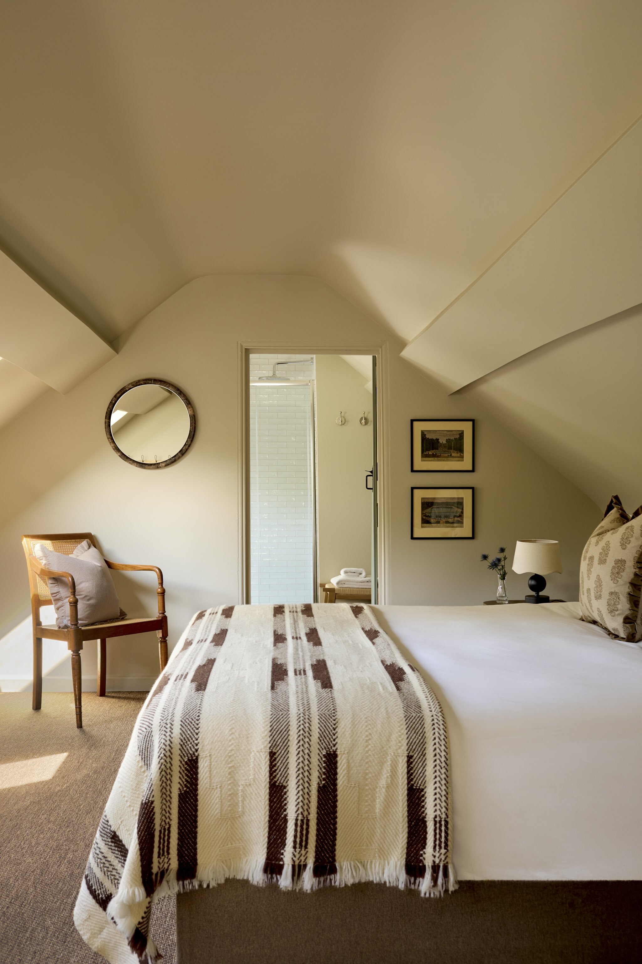 A cozy bedroom with a white bed covered with a brown and white striped blanket, a wooden chair with a beige pillow, a round mirror on the wall, framed artwork, a small table with a lamp and flowers, and a doorway leading to a bathroom with white tile