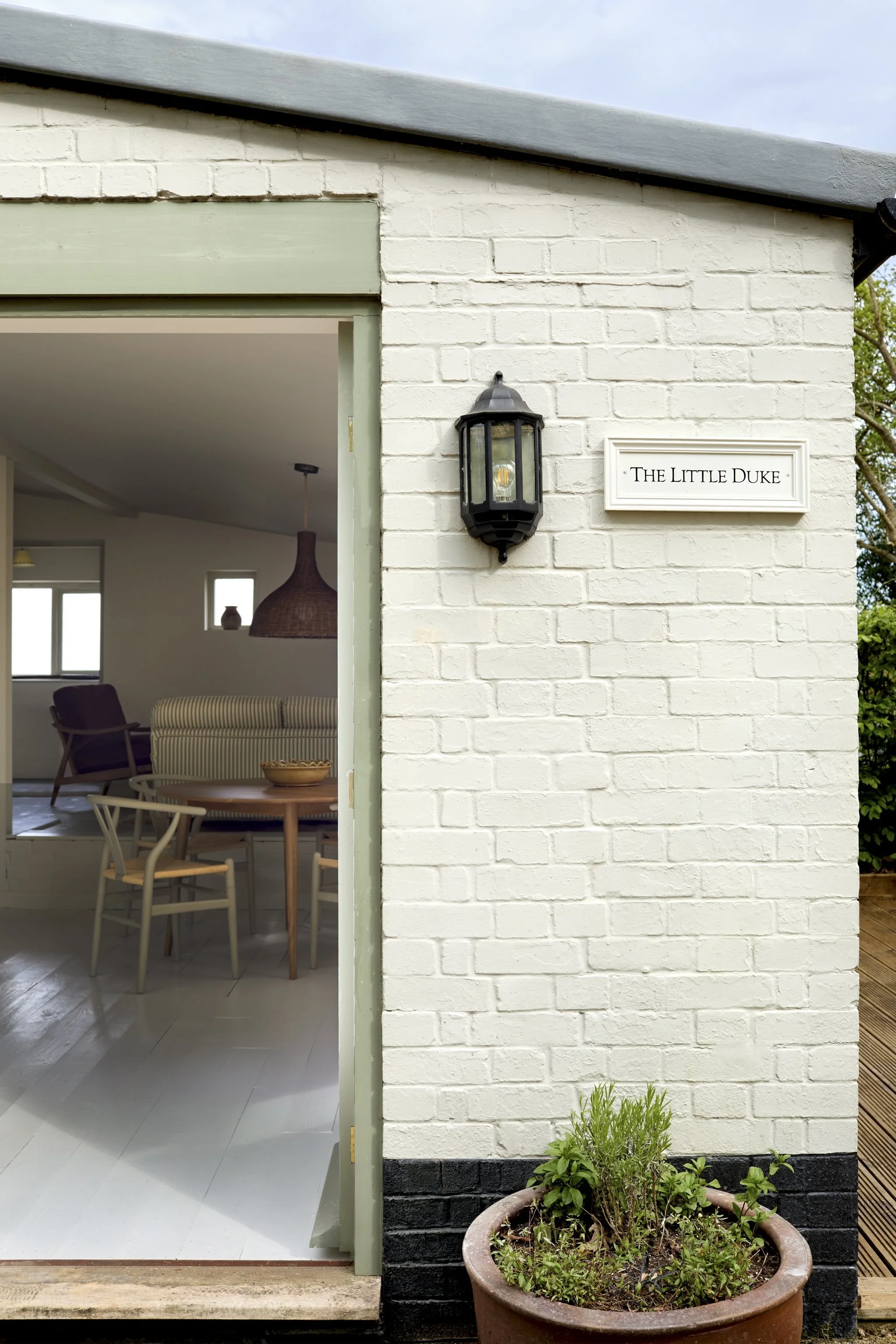 Exterior view of a house showing a white brick wall, a black outdoor wall lantern, a sign reading 'The Little Duke', a flowerpot with plants, and part of an open interior with white furniture and a wicker pendant light