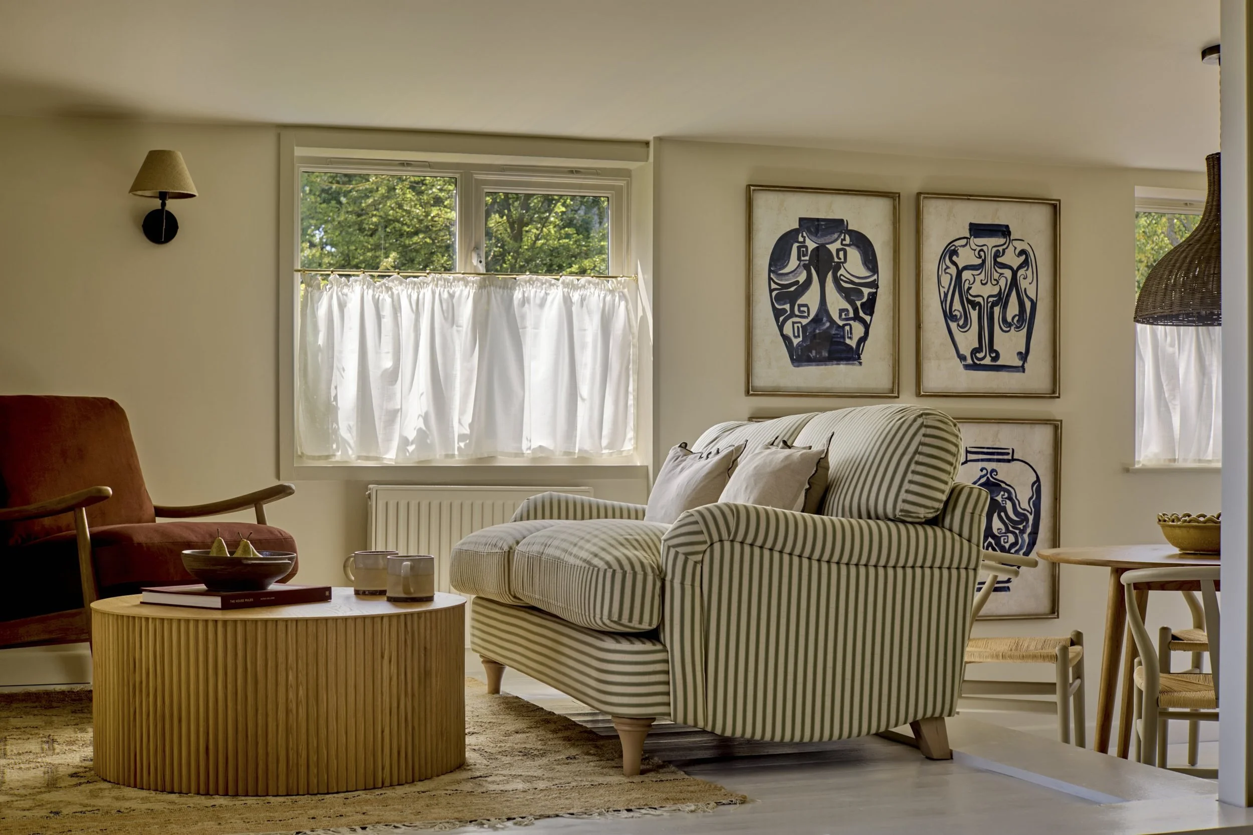 Living room with cream walls, window with white curtains, striped armchair, orange armchair, round wooden coffee table, wall-mounted lamps, framed art, and a woven pendant light.