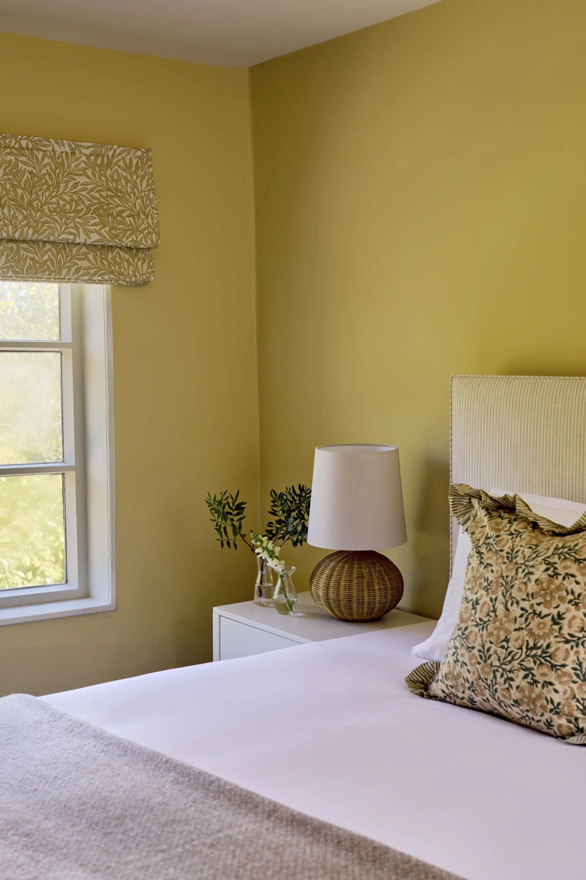 A cozy bedroom with yellow walls, a white bed with a floral pillow, a nightstand with a lamp and a glass vase with green foliage, a window with a botanical patterned curtain, and a rattan lamp base.
