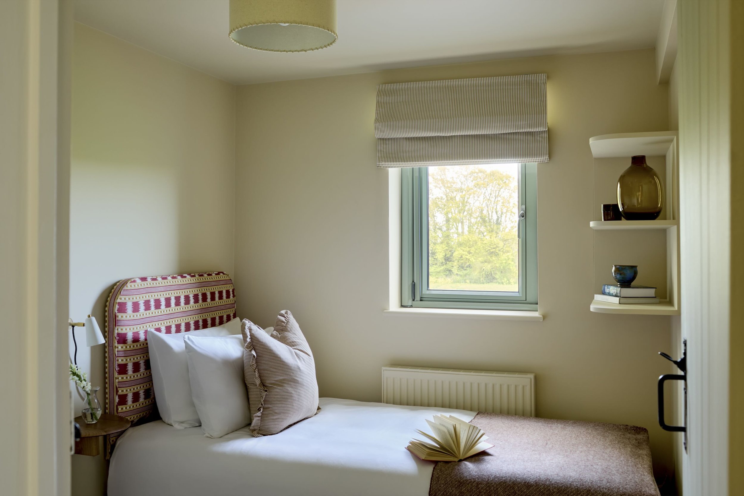 A cozy bedroom with a bed featuring a striped headboard, white bedding, and pillows. There is a window with a striped shade letting in natural light. A floating shelf holds a dark vase and other decorative items. A book is open on the bed with an ope