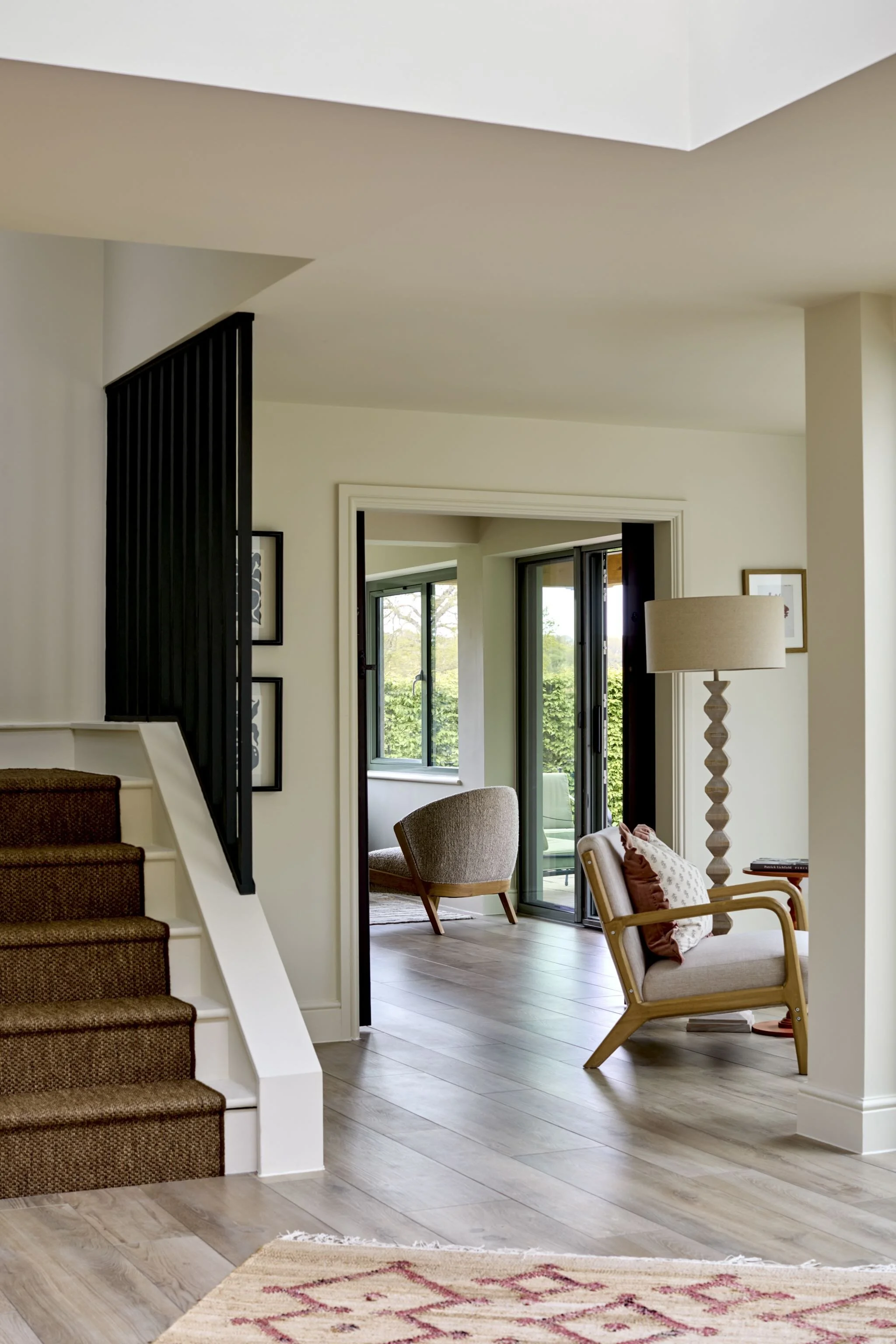 Living room with chairs, floor lamp, windows, and sliding door leading outside. Staircase with brown carpeted steps and black railing visible on the left.