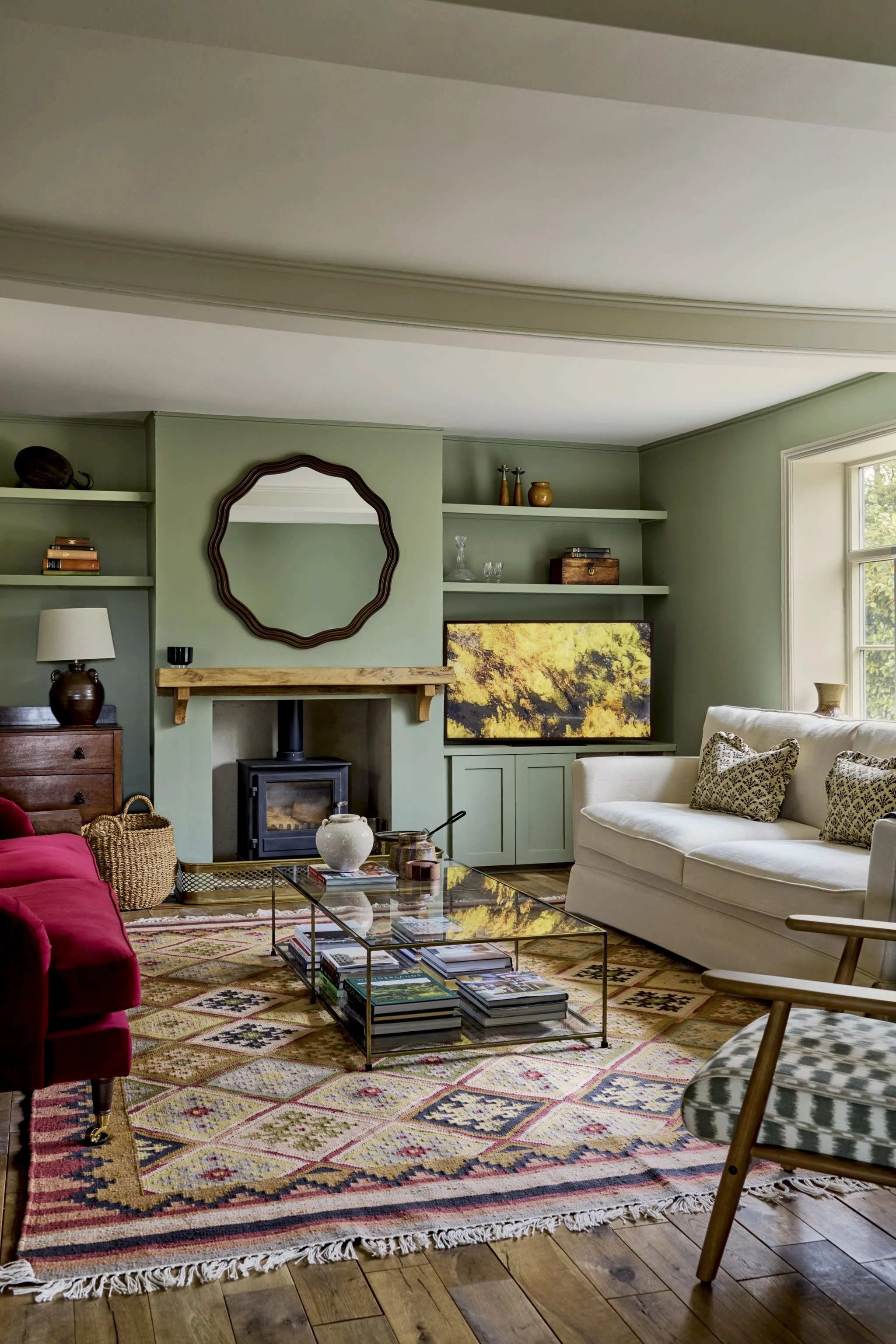 Living room with green walls, a large patterned area rug, a white sofa with patterned pillows, a red velvet chair, a glass coffee table with magazines, a fireplace, a wall-mounted TV, open shelves with decorative items, and a window with sunlight.