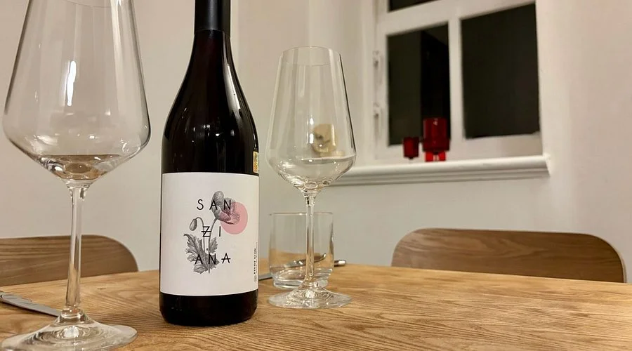 A wine bottle with a white label, two empty wine glasses, a small glass, and a phone on a wooden table in a room with a window and two chairs.