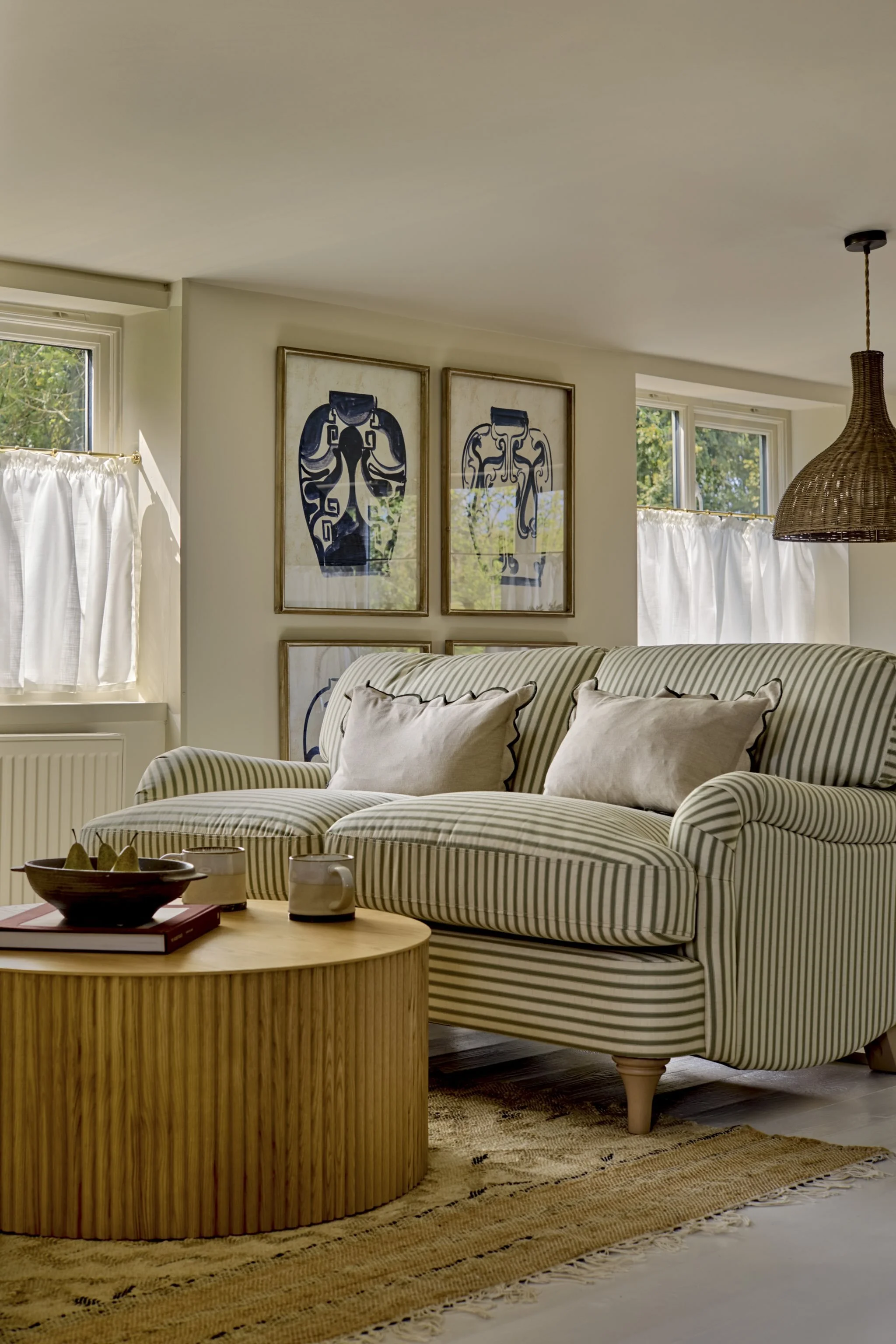 Living room with striped sofa, coffee table with bowls and mugs, framed art on wall, and large windows with white curtains, with a woven pendant light hanging from the ceiling.