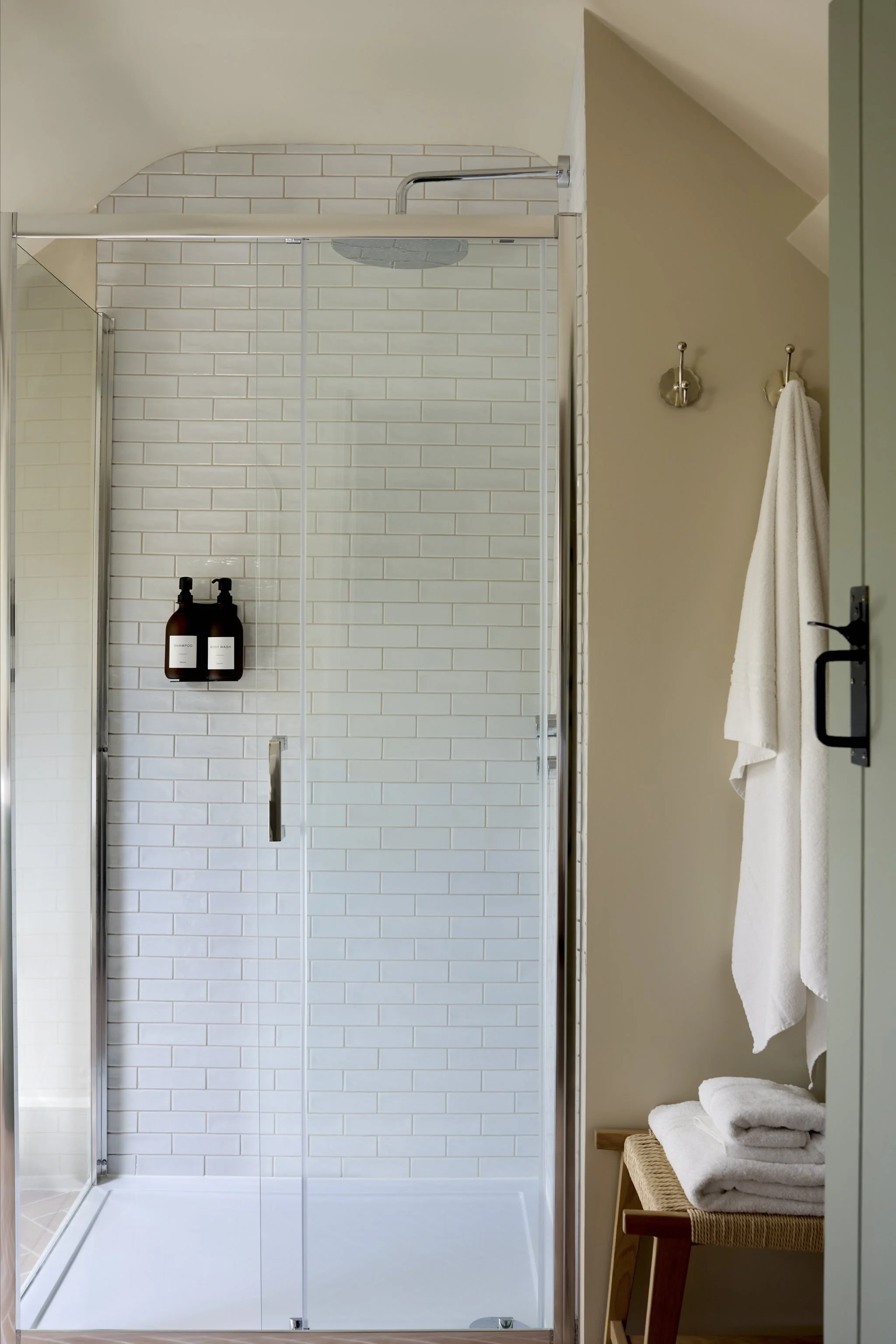 A glass shower enclosure with white subway tiles, a shower head, two brown bottles, and a towel hanging on a wall hook, with towels on a bench beside the shower.