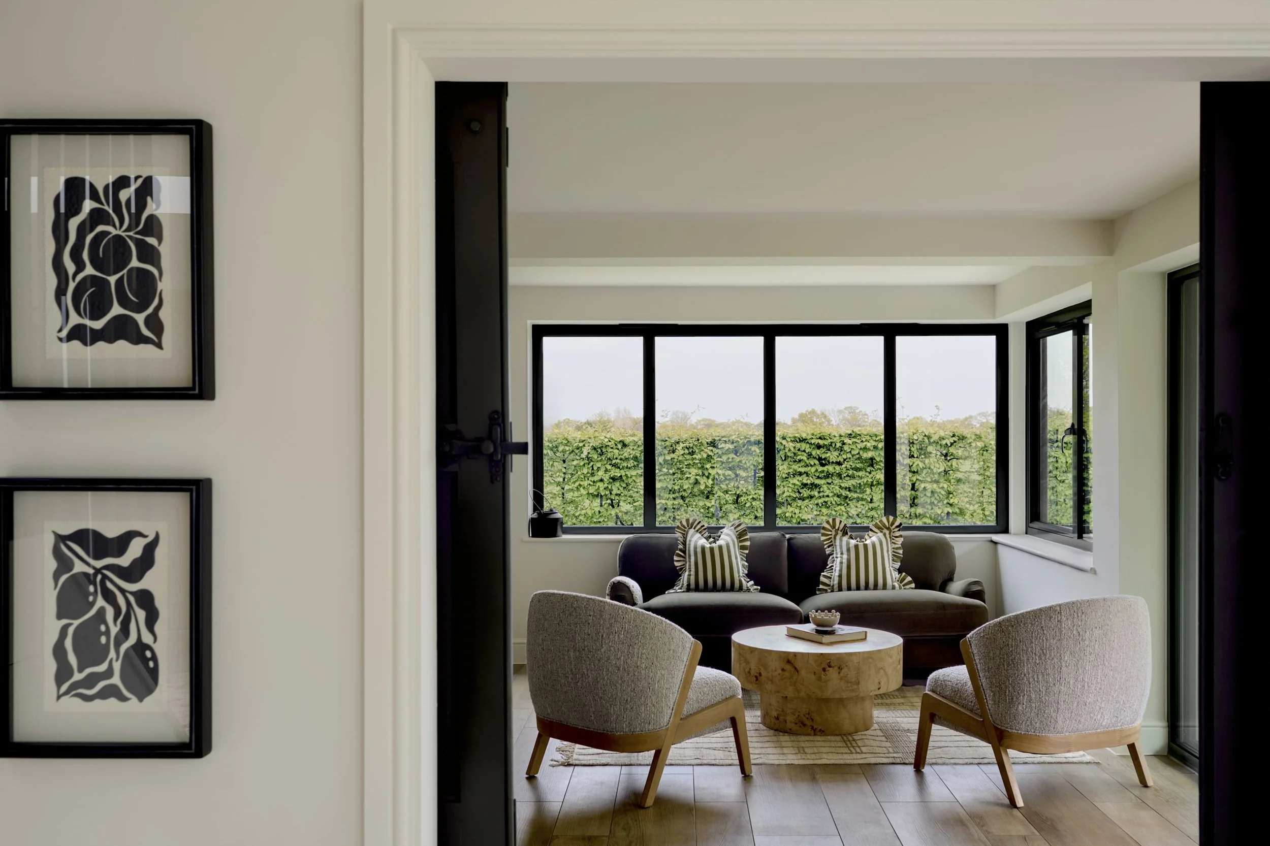 Living room viewed through a doorway, with a large window showcasing green trees outside. The room has a dark sofa with striped pillows, two armchairs, a round wooden coffee table, and framed floral artwork on the wall.