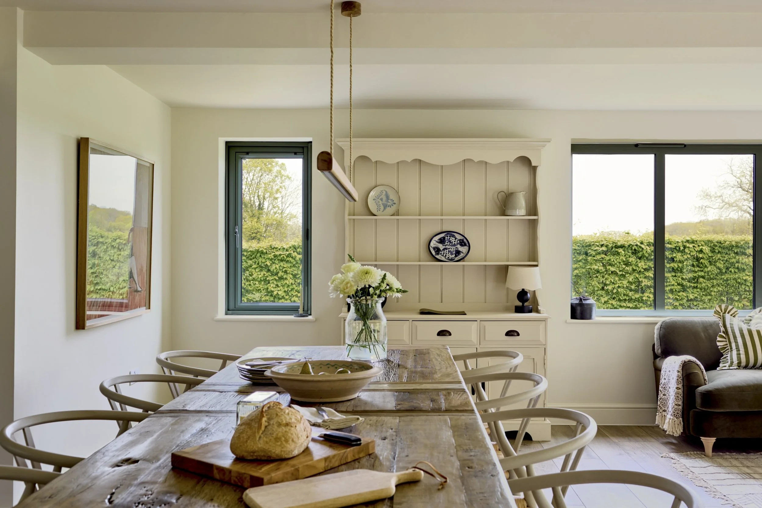 Interior of a dining room with a rustic wooden table set with a bread loaf and a vase of white flowers. The room has large windows showing green trees outside, and a white hutch with decorative plates and a pitcher. There are beige walls and a hangin