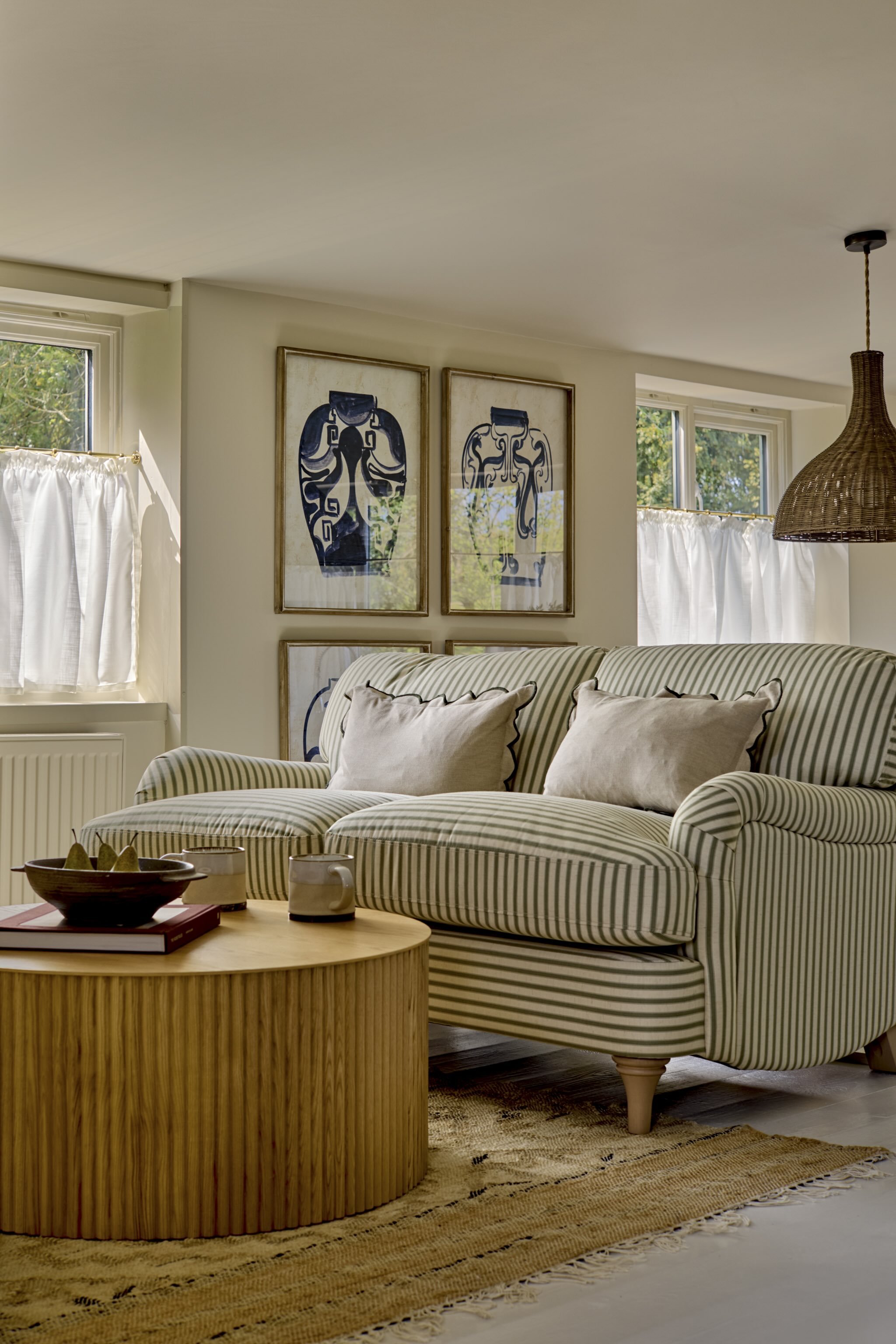 Living room with striped beige and white sofa, pillows, a wooden coffee table with books and bowls, framed artwork of abstract blue figures on the wall, windows with white curtains, woven pendant light, and a patterned rug.