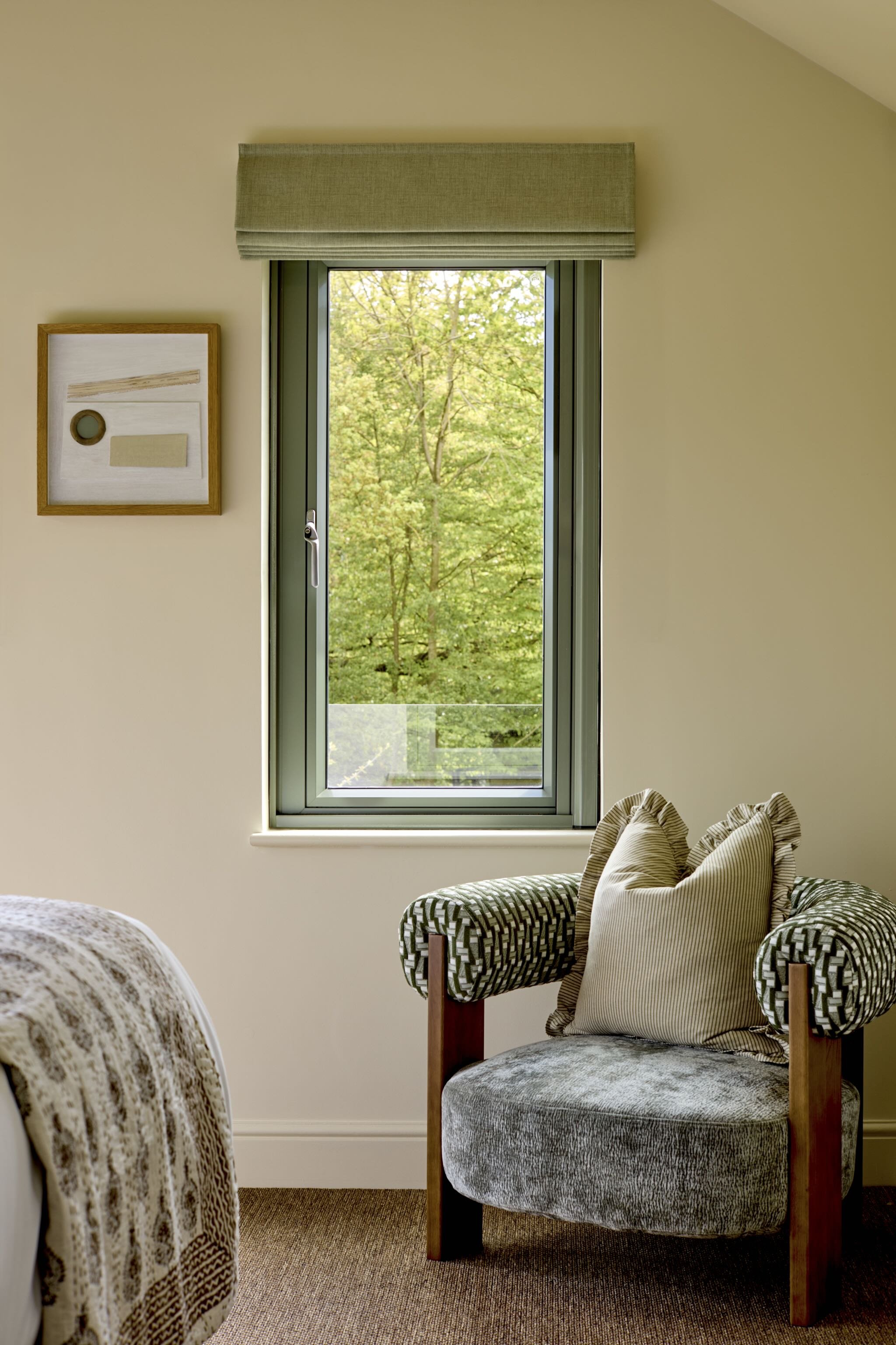 A cozy corner of a room with a large window showing green trees outside. There is a patterned armchair with striped and solid pillows, a textured round ottoman, and artwork on the wall.