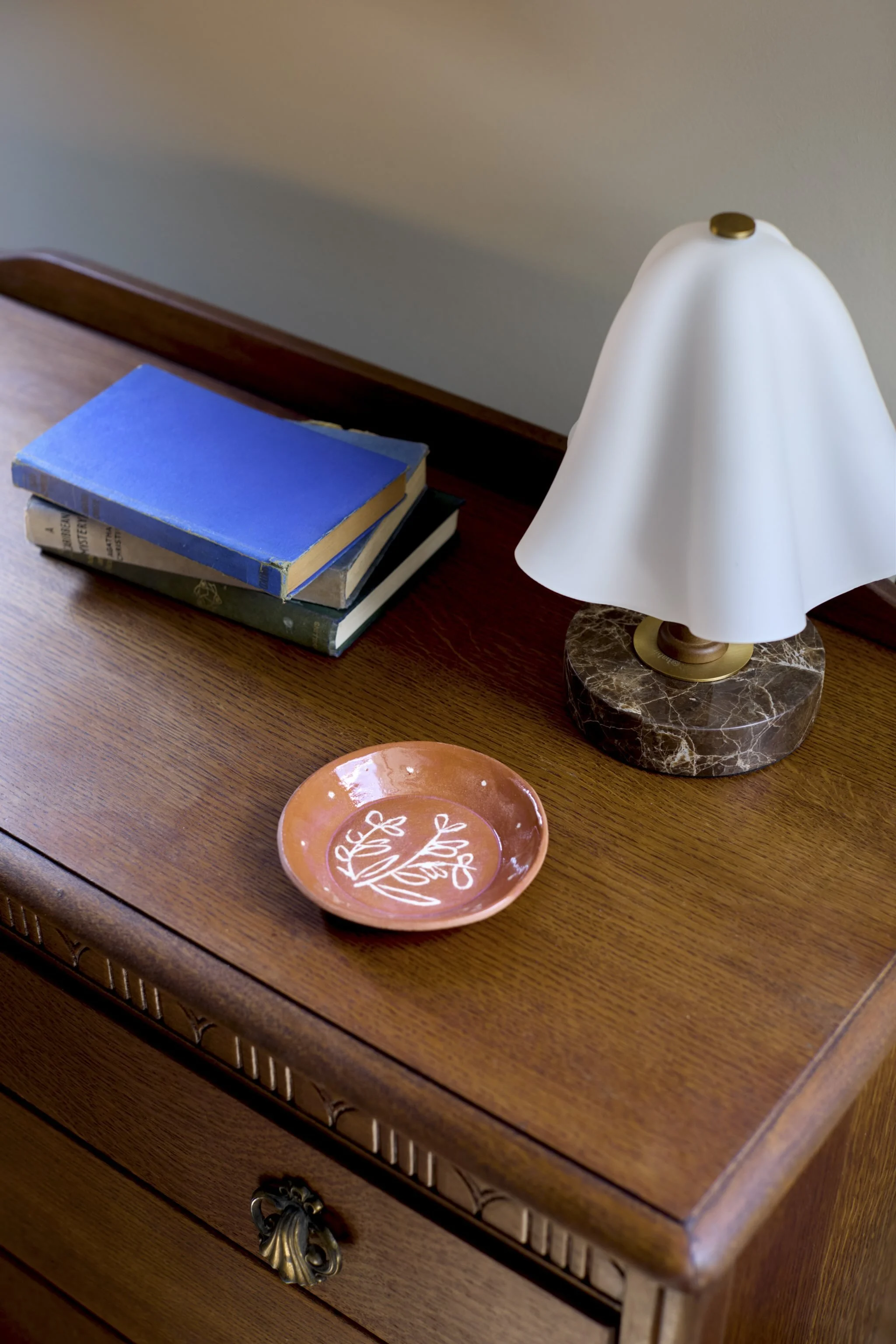 A wooden dresser with a stack of books, a small decorative pink bowl, and a white table lamp with a marble base.