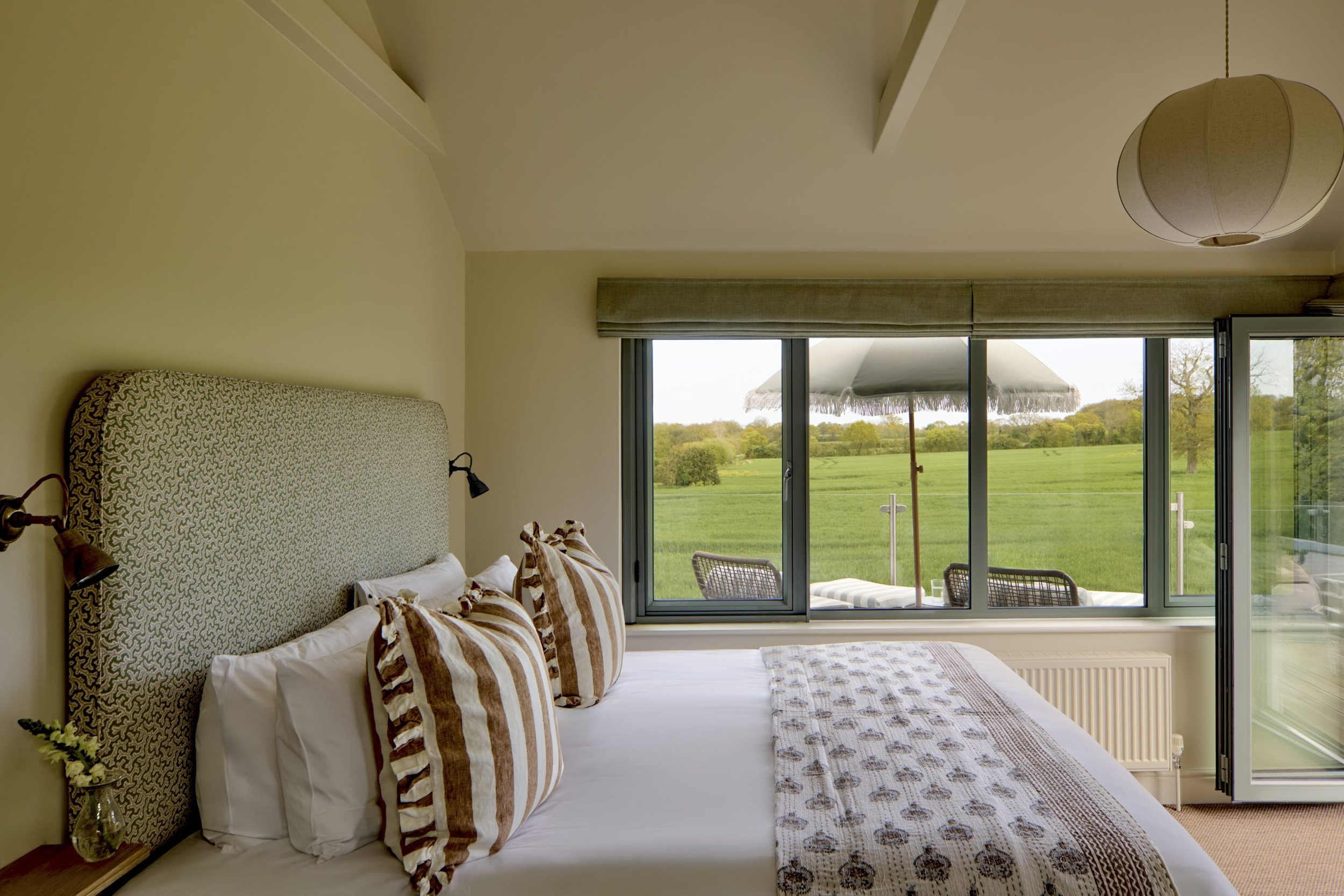 A bedroom with a bed that has a green patterned headboard, striped pillows, and a long patterned blanket. There are black wall-mounted reading lamps and a large window showing a green landscape with trees and a white umbrella outside.