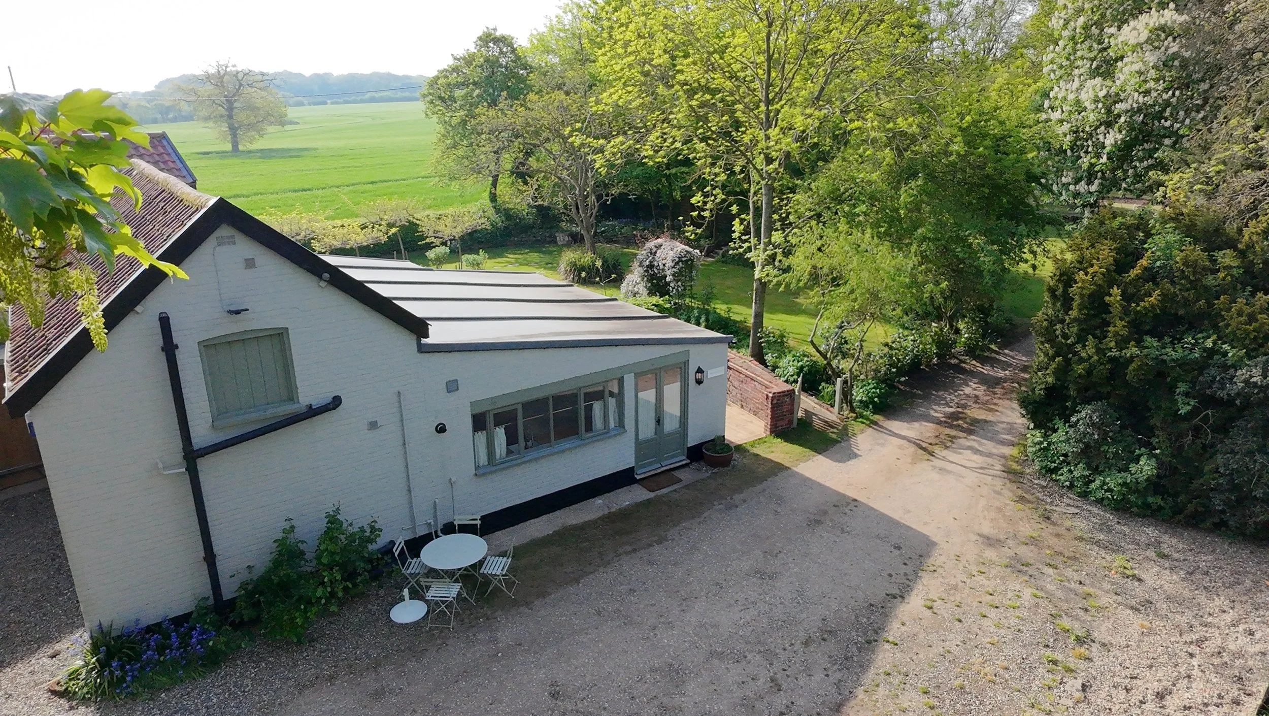 A white house with a black roof, large windows, and a small outdoor seating area with a table and chairs, surrounded by green trees and a gravel path in a rural landscape.