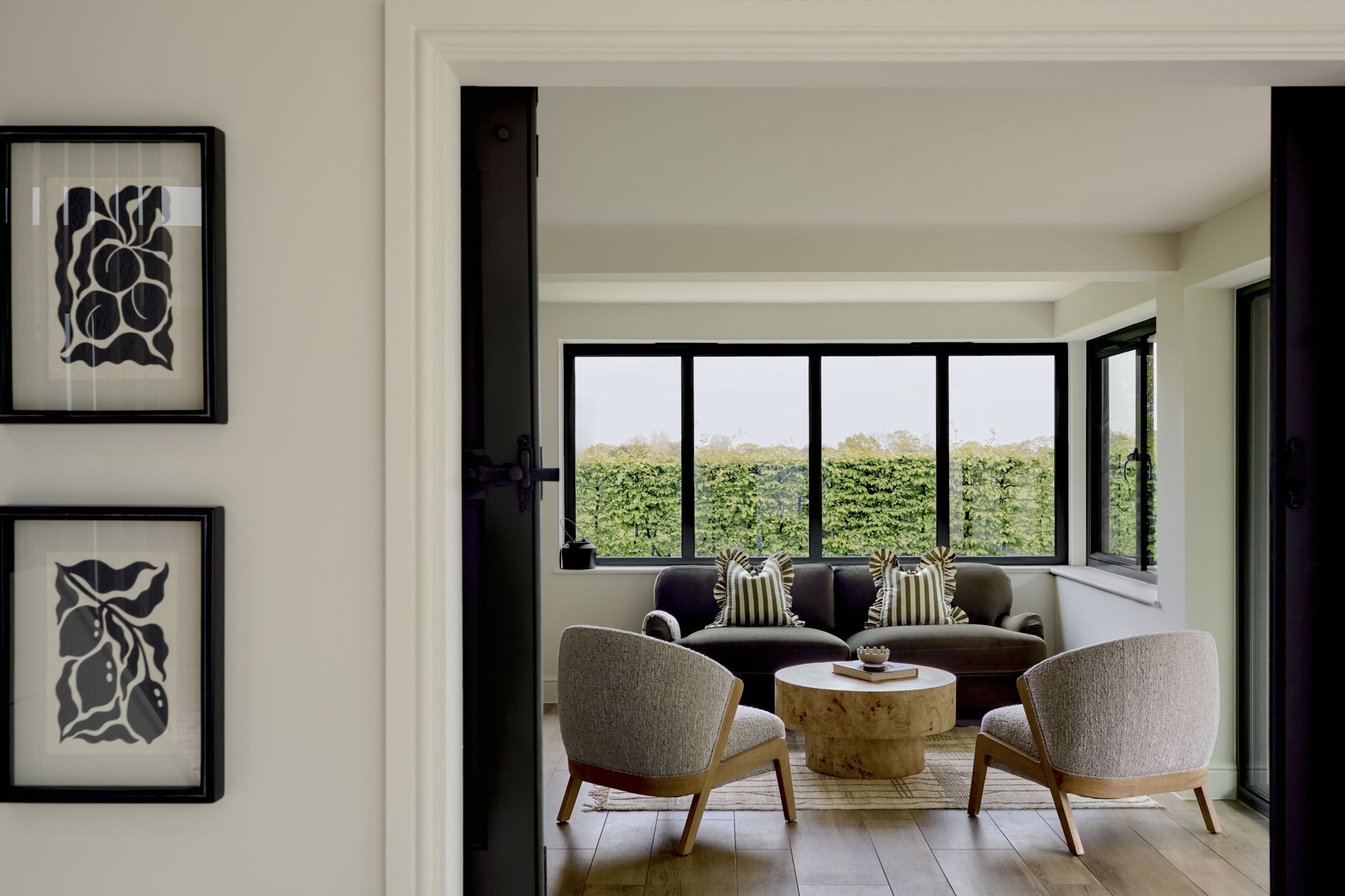 Living room with large window, beige sofa and armchairs, round wooden coffee table, and wall art with black abstract floral design.