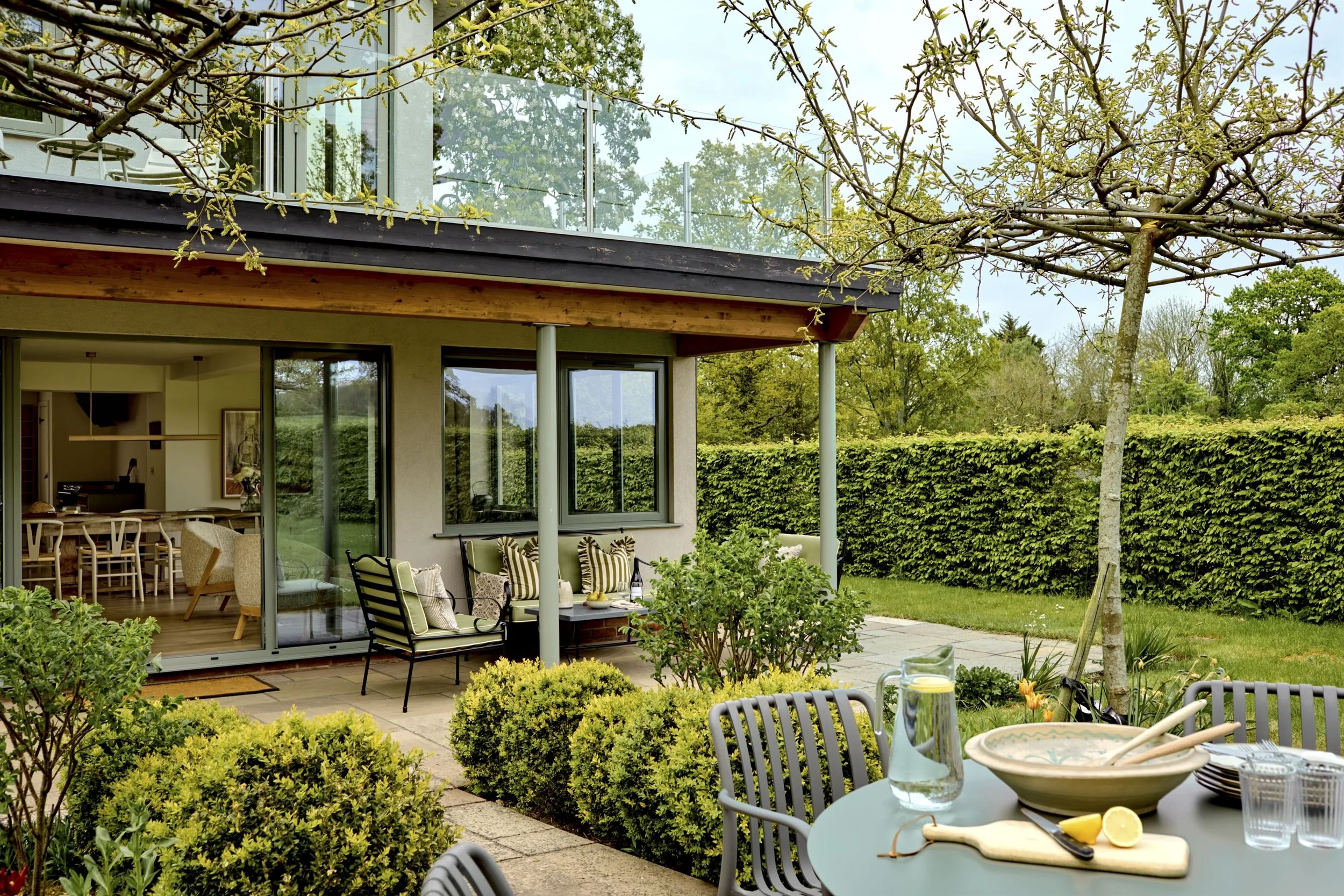 Outdoor patio with dining table, chairs, and glass of water with lemon, surrounded by lush greenery and trees, with a modern house featuring large glass windows and a balcony.
