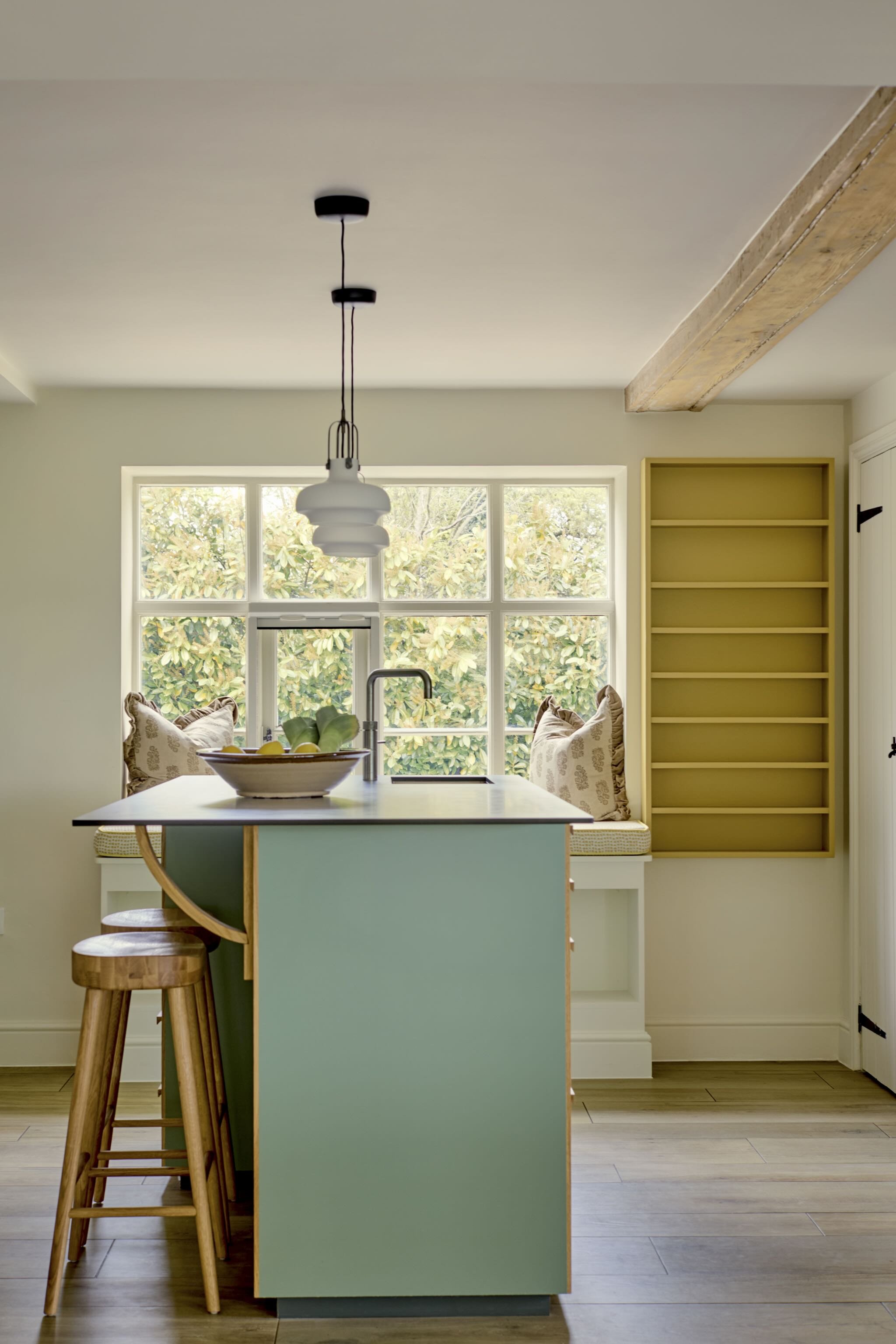 A modern kitchen with a light blue island, wooden stools, a bowl of lemons, a window with greenery outside, yellow shelves, and a pendant light.