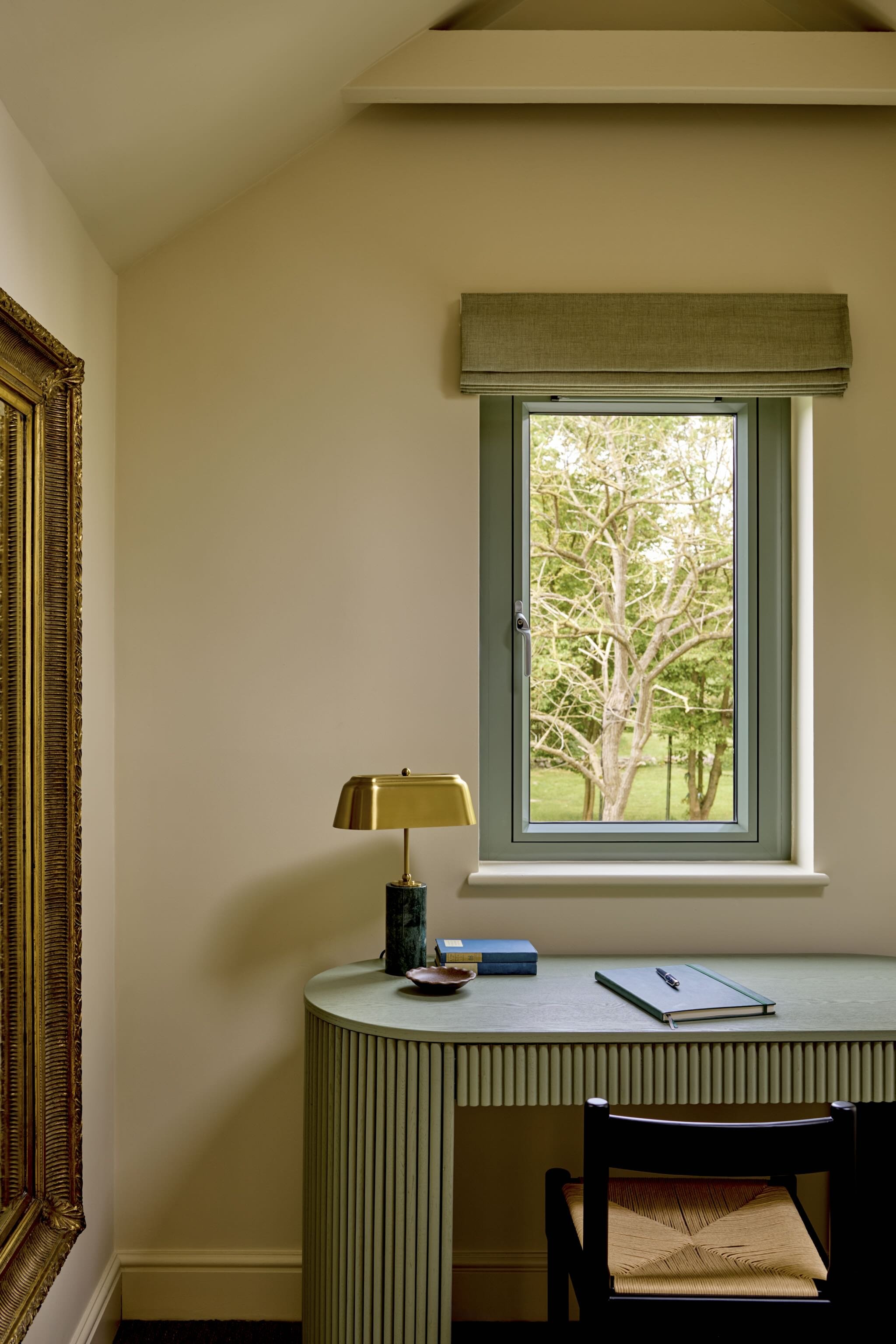 A home office corner with a window showing a tree outside. There is a small desk with a gold lamp, blue books, a notepad, and pen. A black chair with a woven seat is in front of the desk. Part of a large gold-framed mirror or artwork is visible on th