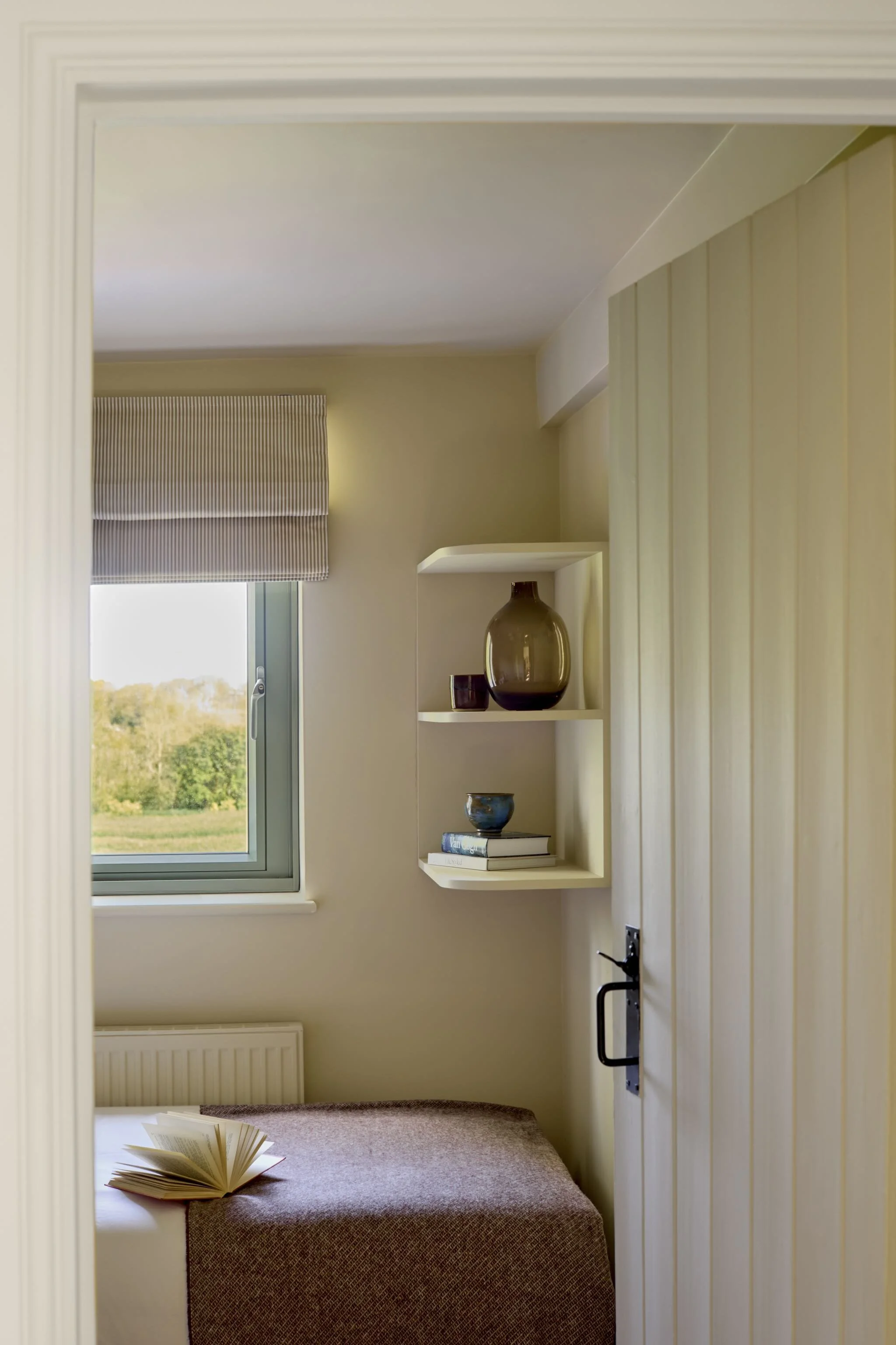 A cozy bedroom with a window covered by a gray and white striped curtain, a small white shelf with decorative items, a brown upholstered bed with an open book on it, beige walls, and a neutral carpeted floor.
