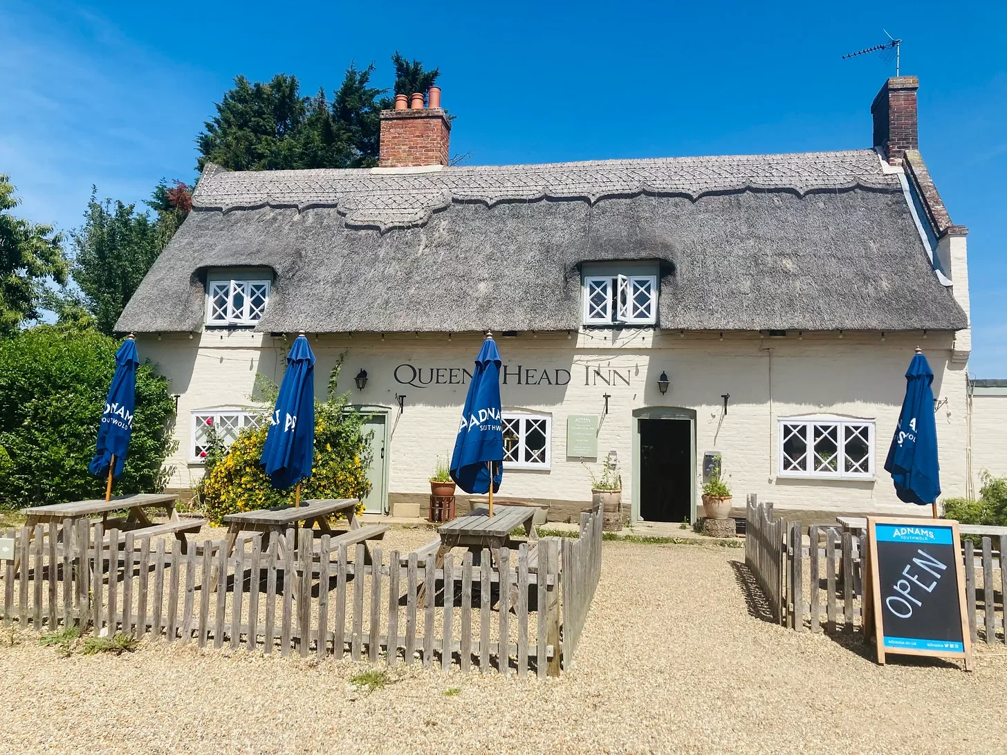 Exterior of a traditional English pub named Queen Head Inn with outdoor seating area, blue umbrellas, and a chalkboard sign that says open.