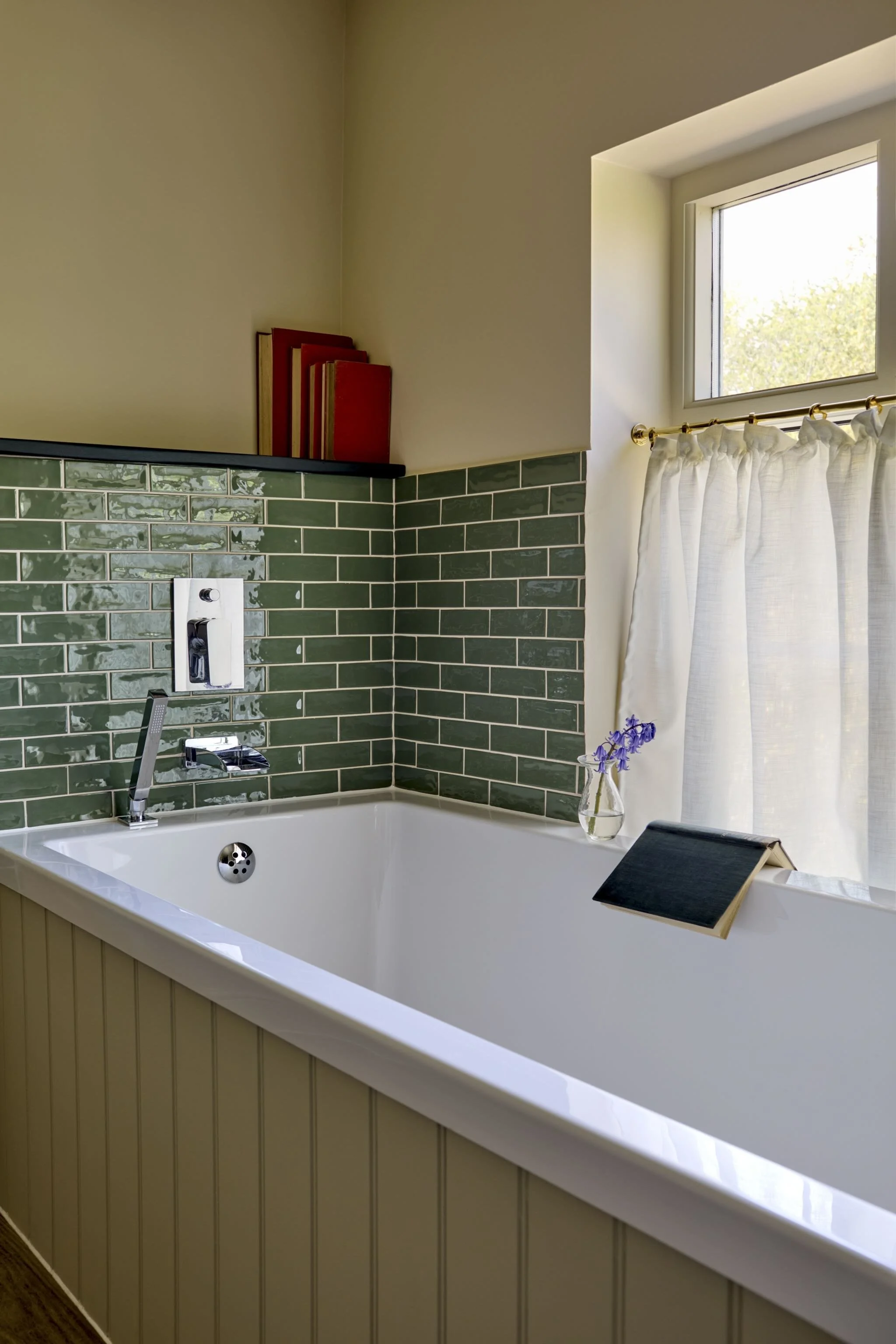A bathroom with a white bathtub, green subway tile backsplash, a window with white curtain, and a small table with a black slat in front of the tub. There are red books on a shelf and a vase with purple flowers inside the tub.