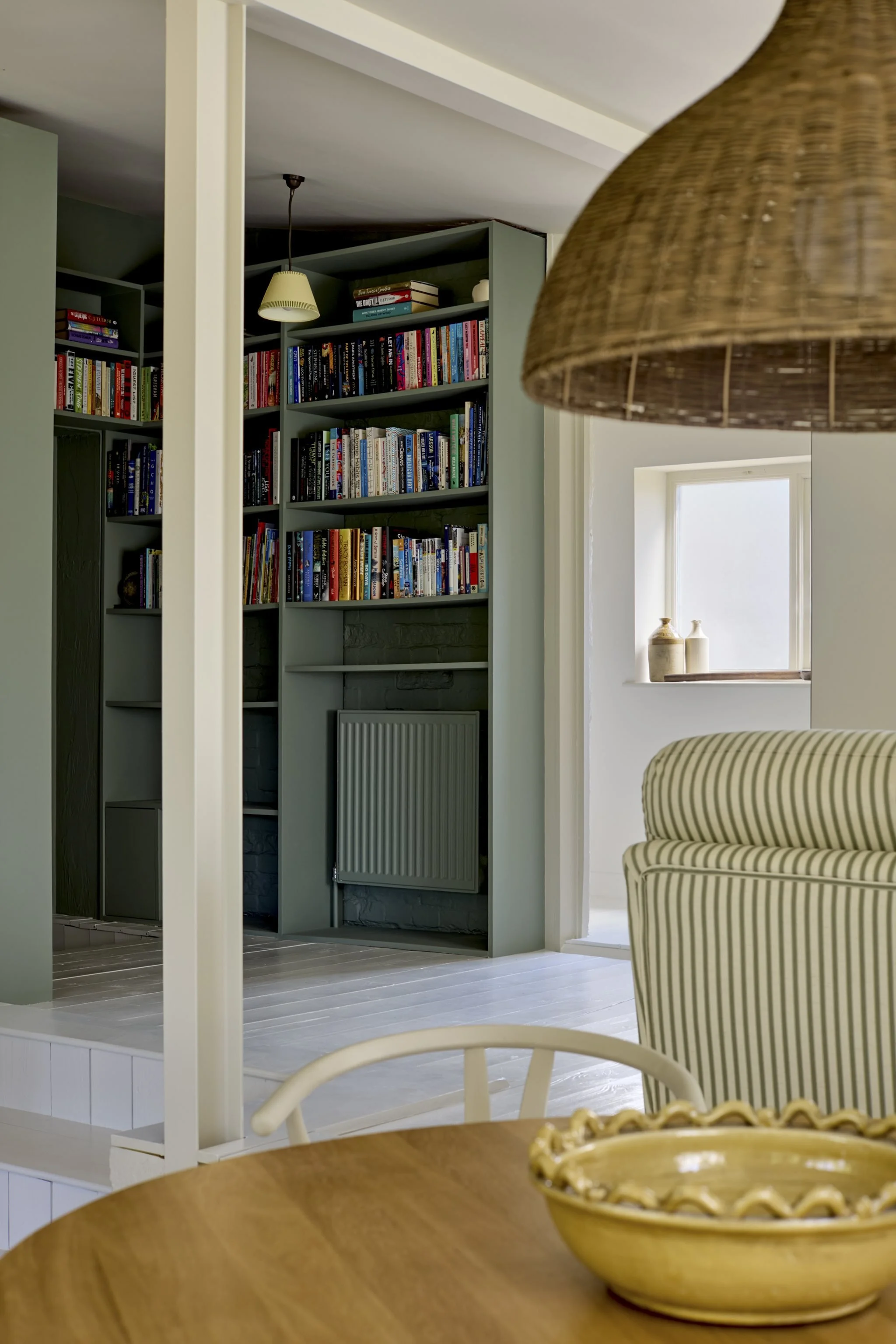 A cozy living space with a round wooden dining table in the foreground, a striped armchair, and a large wicker lampshade hanging from the ceiling. In the background, there is a bookshelf filled with books, a small window with decorative pottery, and 